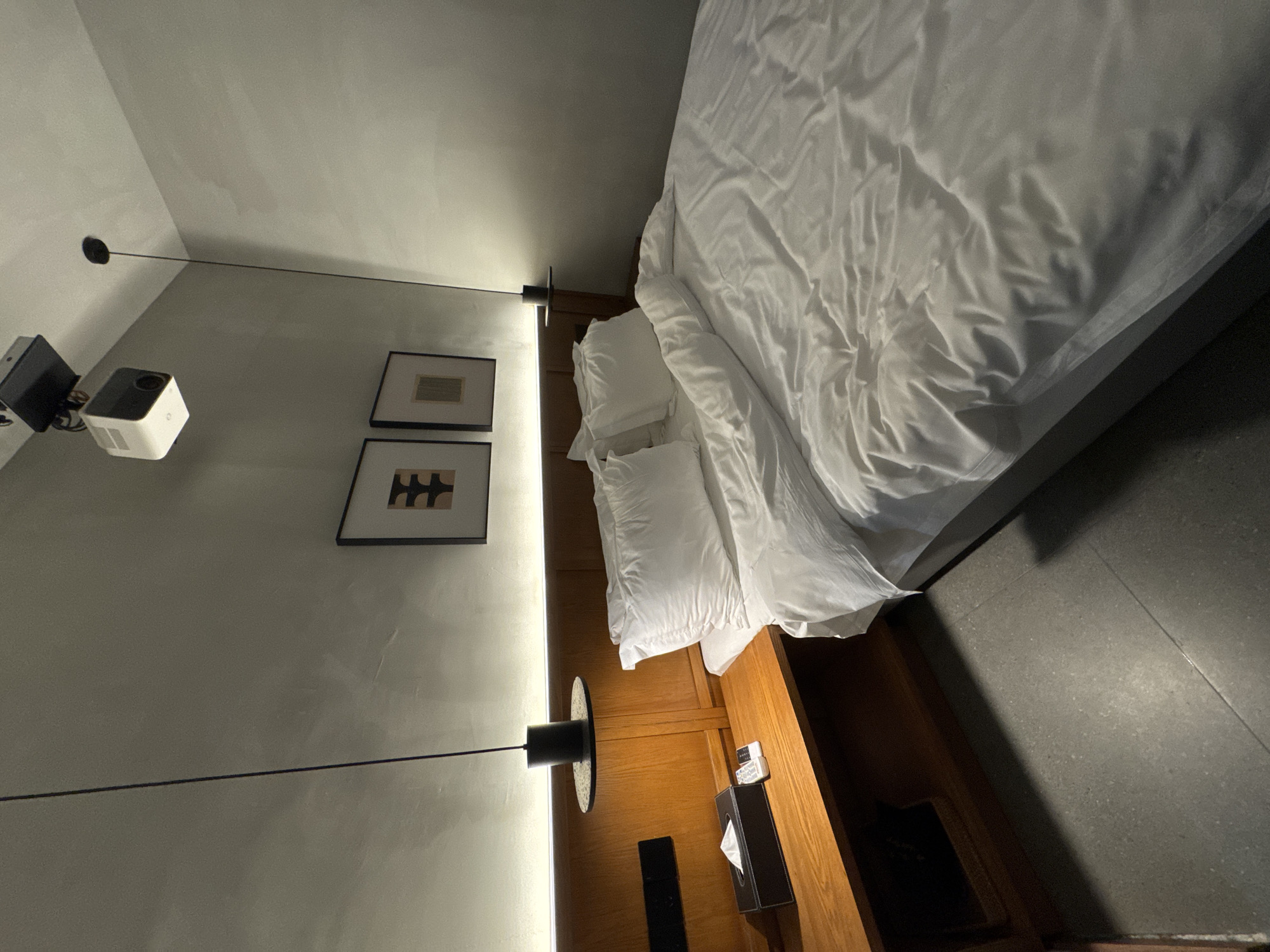 The image presents an interior scene of what appears to be a hotel room or bedroom. The composition is dominated by a large bed dressed in white linens. Two pillows are positioned at the head of the bed. To the left of the bed, there is a wooden headboard with a built-in shelf or nightstand. On the nightstand, there is a tissue box and some small items. On the wall, there are two framed pieces of artwork. A lighting fixture, seemingly a hanging lamp, is visible on the left side of the image, with the cable extending towards a projector and possibly an associated black box, also mounted on the wall. The walls are light grey. The floor appears to be a dark grey tile. The location is given as Wulong District, China, but no further information to confirm this is present in the image. No people are visible in the image.