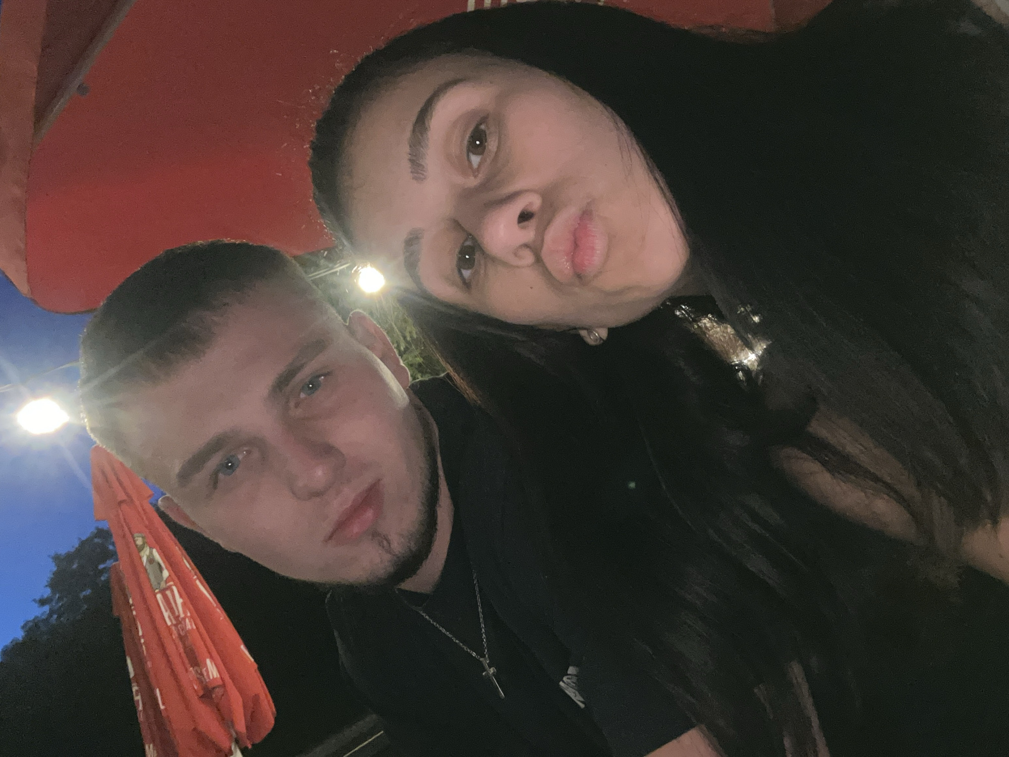 Two individuals, a male and a female, are captured in a close-up selfie outdoors. The male, on the left, has short, light brown hair, blue eyes, and a light beard with a goatee. He is wearing a black t-shirt with a silver cross necklace. The female, on the right, has dark, long hair and is making a kissing face. She appears to be wearing a dark hoodie. Behind them, large red umbrellas are visible, suggesting they might be at an outdoor seating area of a restaurant or cafe. The sky is dark blue with a bright light source visible in the upper left quadrant, possibly a streetlamp. The scene is illuminated by artificial light, casting shadows and creating a dimly lit atmosphere. The background suggests an urban environment at night.