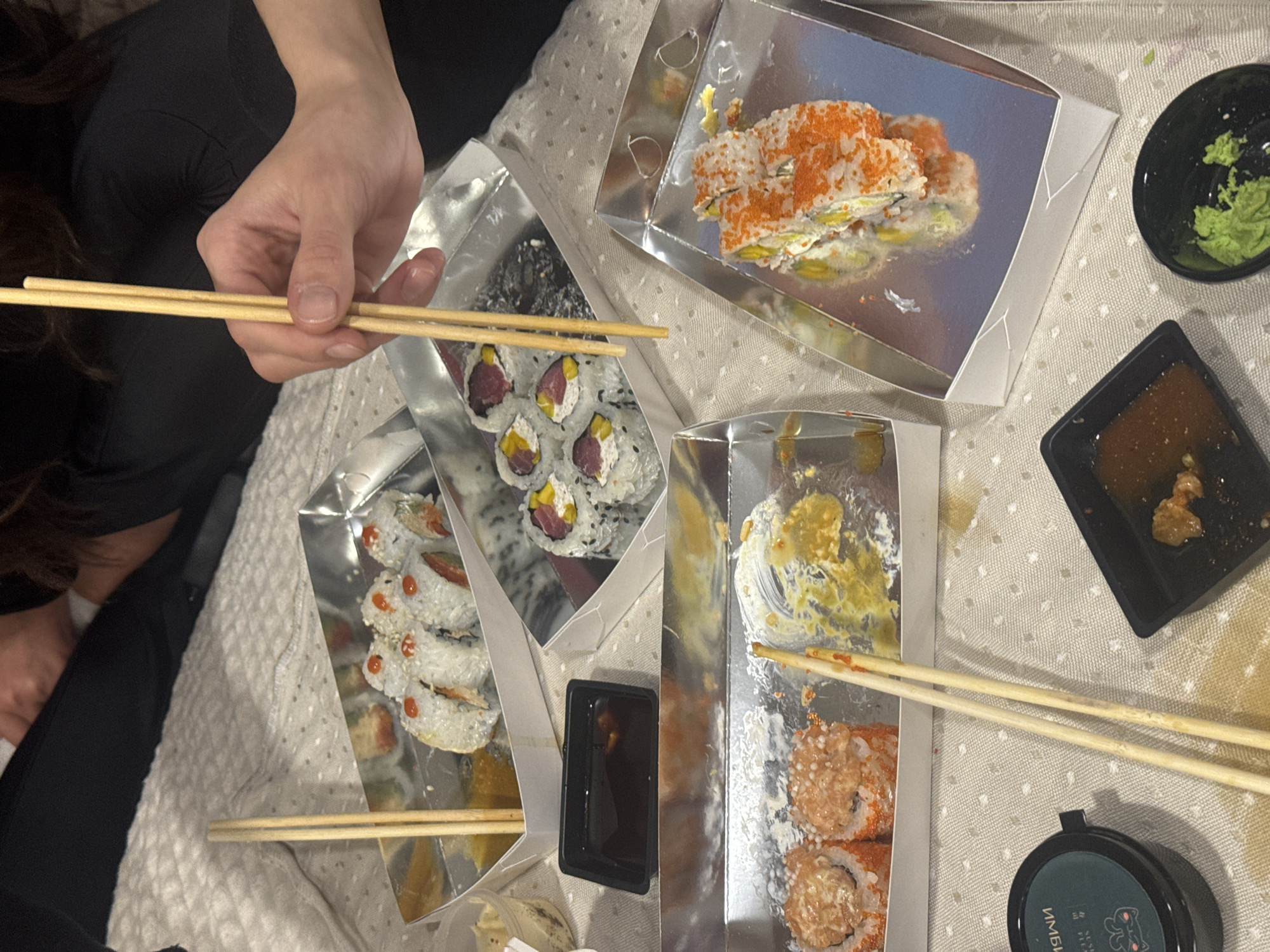 An overhead shot captures a casual sushi meal for at least two people, likely indoors in Krasnoyarsk, Russia. Two hands are visible; one holds a pair of wooden chopsticks over a box of sushi, while another pair of chopsticks rests in a different container.
Several types of sushi rolls are arranged in rectangular, silver-lined cardboard takeaway boxes. One box contains rolls topped with bright orange fish roe. Another features rolls with dark sesame seeds, pieces of tuna, and a yellow filling, possibly mango or a similar ingredient. A third box holds white rice rolls adorned with a red sauce drizzle and sesame seeds. A fourth partially eaten box shows remnants of a yellow sauce.
Various condiments accompany the meal. A small black bowl in the upper right corner contains a serving of bright green wasabi. Below it, a square black dish holds a dark brown liquid, presumably soy sauce, with some solid pieces. At the bottom right, a small round black container is visible, featuring the Russian word "ИМБИРЬ" (meaning "GINGER") and a small cat logo on its label. The entire setup rests on a light-colored, subtly patterned textile surface, possibly a tablecloth or blanket, suggesting a relaxed dining atmosphere.