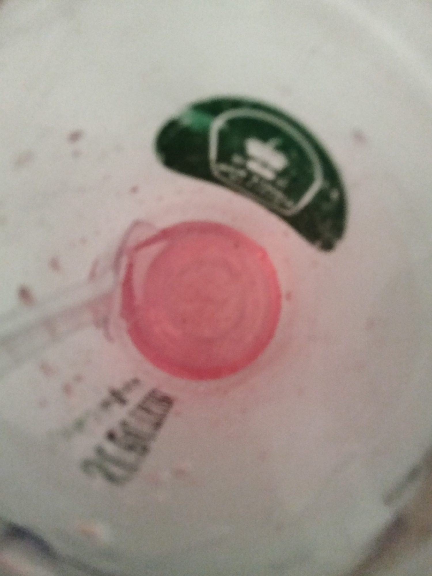 The image shows a top-down view of a pink-colored object, possibly a cap or lid, inside a container. A green label with white text and a white outline is partially visible in the upper right quadrant of the image. The label appears to be a seal or branding sticker. Some faint pinkish splotches are scattered across the white background of the container. In the lower left quadrant, some black text is printed or stamped on the container, appearing to be a product code or batch number, partially obscured by the pink object. The lighting is somewhat dim, creating a shallow depth of field. The image provides no information to determine the location in the city of Payagaboh, Indonesia.