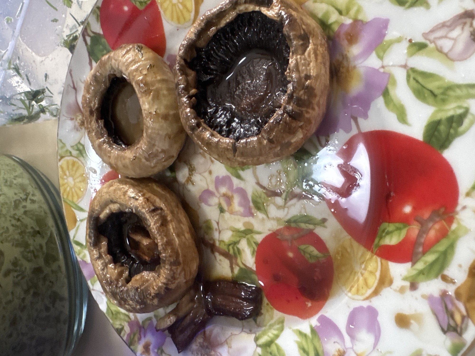 The image depicts a plate containing cooked mushrooms. There are three mushroom caps, two with dark interiors and one with a light interior. A small piece of mushroom stem lies near the bottom of the plate. The plate is decorated with images of red tomatoes, lemon slices, and purple flowers. In the background, on the left side of the image, there is a glass bowl or container with a green substance inside. The location of the image is unknown, and there is no evidence that it was taken in Tbilisi, Georgia.