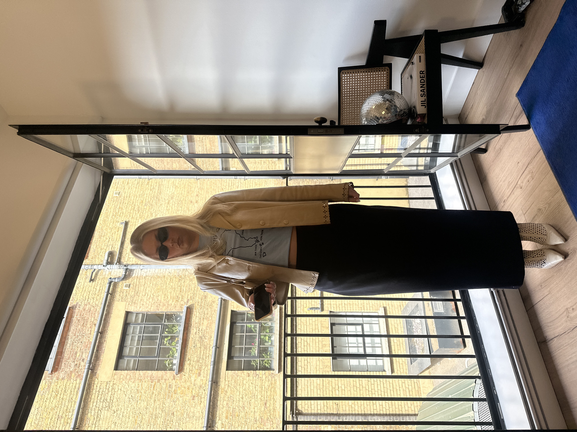 A woman is standing on a balcony in London, United Kingdom. She is wearing sunglasses, a light-colored jacket, a t-shirt, a long black skirt, and white shoes. She is holding a mobile phone. In the background, there is a yellow brick building with windows and pipes. The balcony has a black metal railing. Inside the room, there is a black shelf with a disco ball and a book titled "JIL SANDER". There is also a blue mat on the floor.