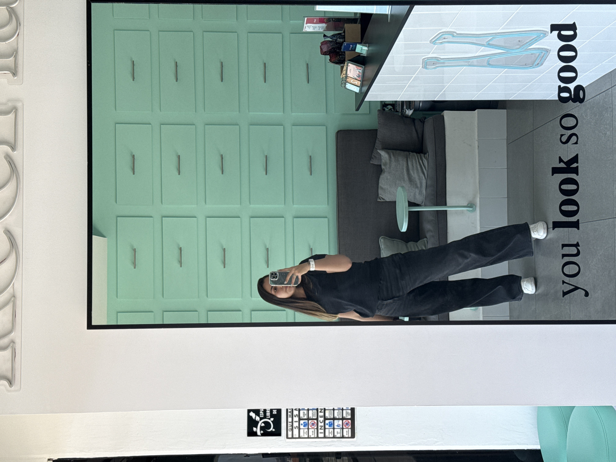 The image captures a full-body mirror selfie of a young woman in an indoor setting. The woman stands upright, holding a smartphone to take the photo, which partially obscures her face. She has long, dark hair and is dressed in a black t-shirt, black wide-leg pants, and white sneakers. A watch is visible on her left wrist.
The reflection in the mirror reveals a room with a distinct aesthetic. The background wall behind the woman is painted a light mint green and features numerous rectangular panels, each fitted with a silver handle, giving the appearance of decorative drawers or cabinet fronts. To the right of the woman in the reflection, a dark-surfaced table holds various items, including small boxes, what appear to be books, and a flip calendar. Below the table, a dark gray sofa with lighter gray cushions and a throw pillow is visible. A slender, light green stand with a circular top extends from the floor near the sofa. The floor within the reflected space is light gray tile.
On the right side of the mirror, oriented vertically, black text reads "you look so good." Above this text, a graphic depicts two stylized, light blue-outlined objects that resemble shoes or feet.
Outside the mirror's frame, on the left, there is a white wall with raised, metallic-looking, stylized shapes that could be letters or numbers. Below the mirror, mounted on a white panel, are two small informational signs. One sign has a black background with a white plant-like icon and partially visible text "ESPA LIBRE DE." The other sign features several small icons and text fragments like "QUE HAC SIS INCEN," suggesting safety instructions, possibly related to fire prevention. The scene is brightly and evenly lit, indicating an indoor environment.