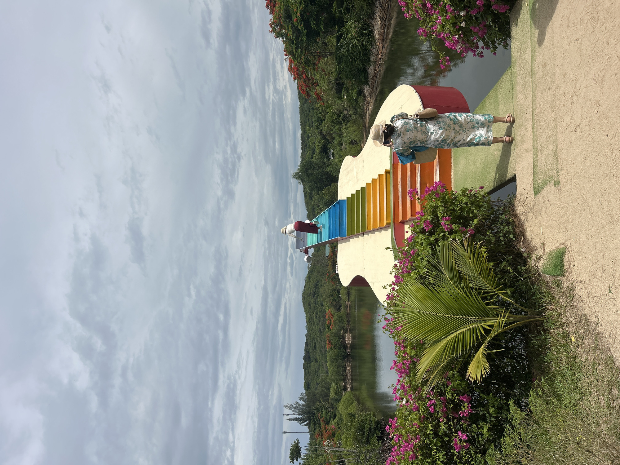 The image depicts an outdoor scene featuring a guitar-shaped structure with a rainbow-colored staircase. Two individuals are visible: one woman wearing a straw hat and a floral dress stands facing away from the camera, holding a camera and a bag, on a sandy pathway. Another person, partially obscured, is ascending the rainbow stairs. The structure is situated next to a body of water, with lush green trees, shrubs, and vibrant pink flowers surrounding the area. In the background, rolling hills covered in greenery are visible under a cloudy sky. The location is identified as Hoa Thang city, Vietnam.
