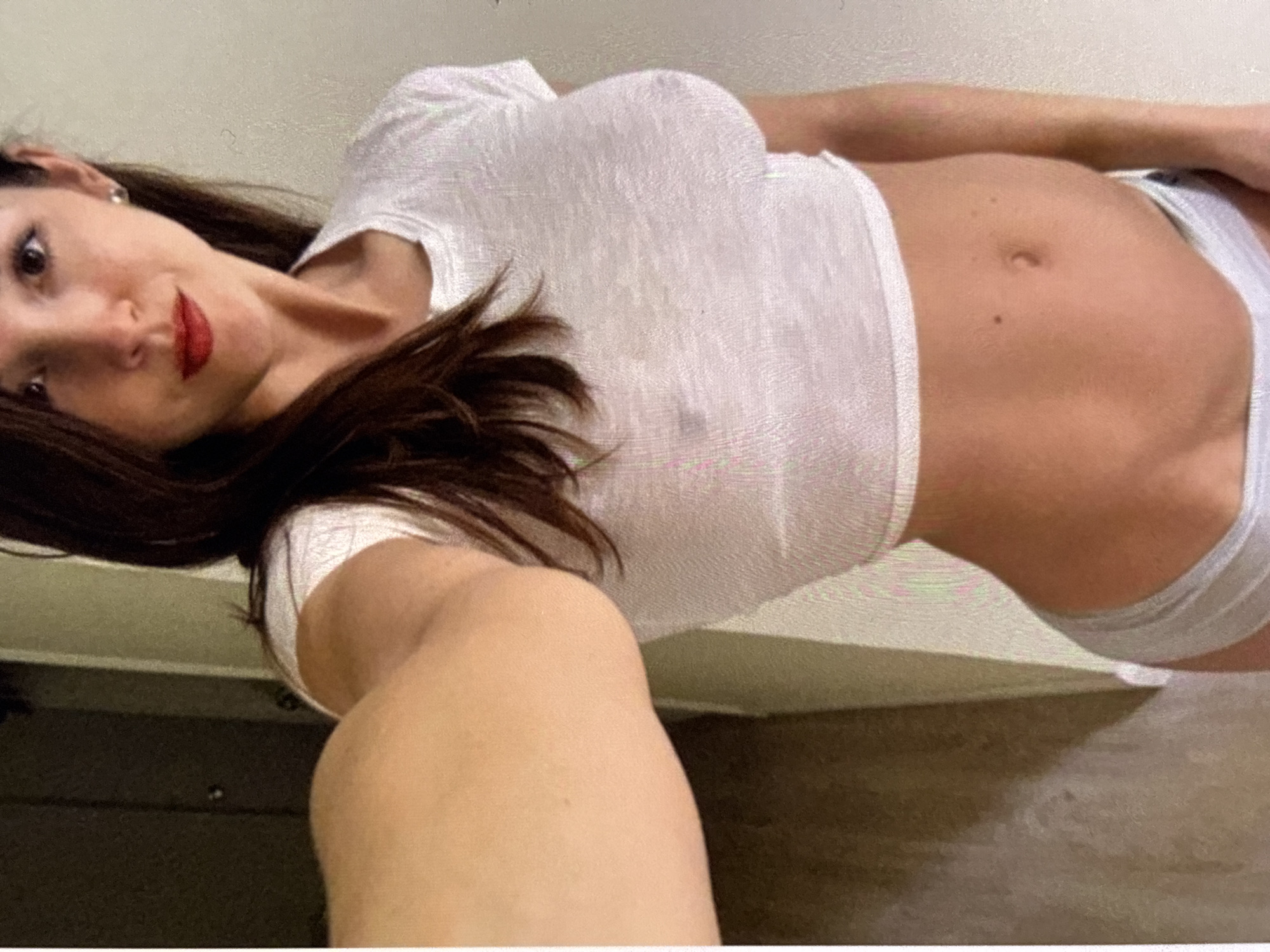 The image shows a woman in a white, sheer crop top and light-colored underwear. She has dark hair, red lipstick, and is wearing a small earring in her right ear. She is taking a selfie with her left arm extended. The background appears to be an indoor setting with a wall and a partially visible doorway. The lighting is soft.
