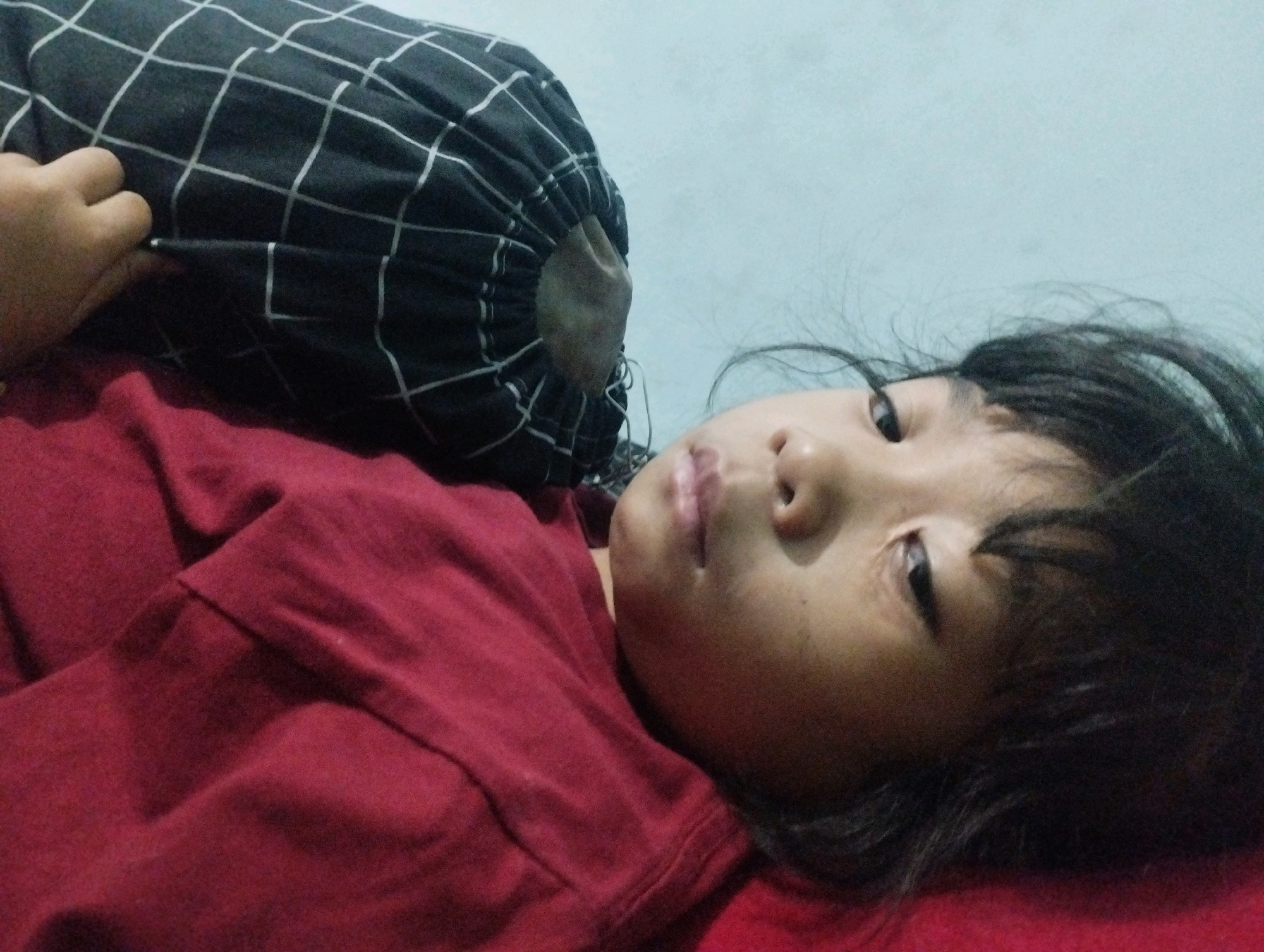 A young child with dark hair is lying on their back on a red surface, possibly a bed, with a pillow featuring a black and white grid pattern behind their head. The child is wearing a dark red shirt. The child's face is partially obscured by their hair and the pillow. The background is a pale blue wall. The image was taken in Kemantren, Indonesia.