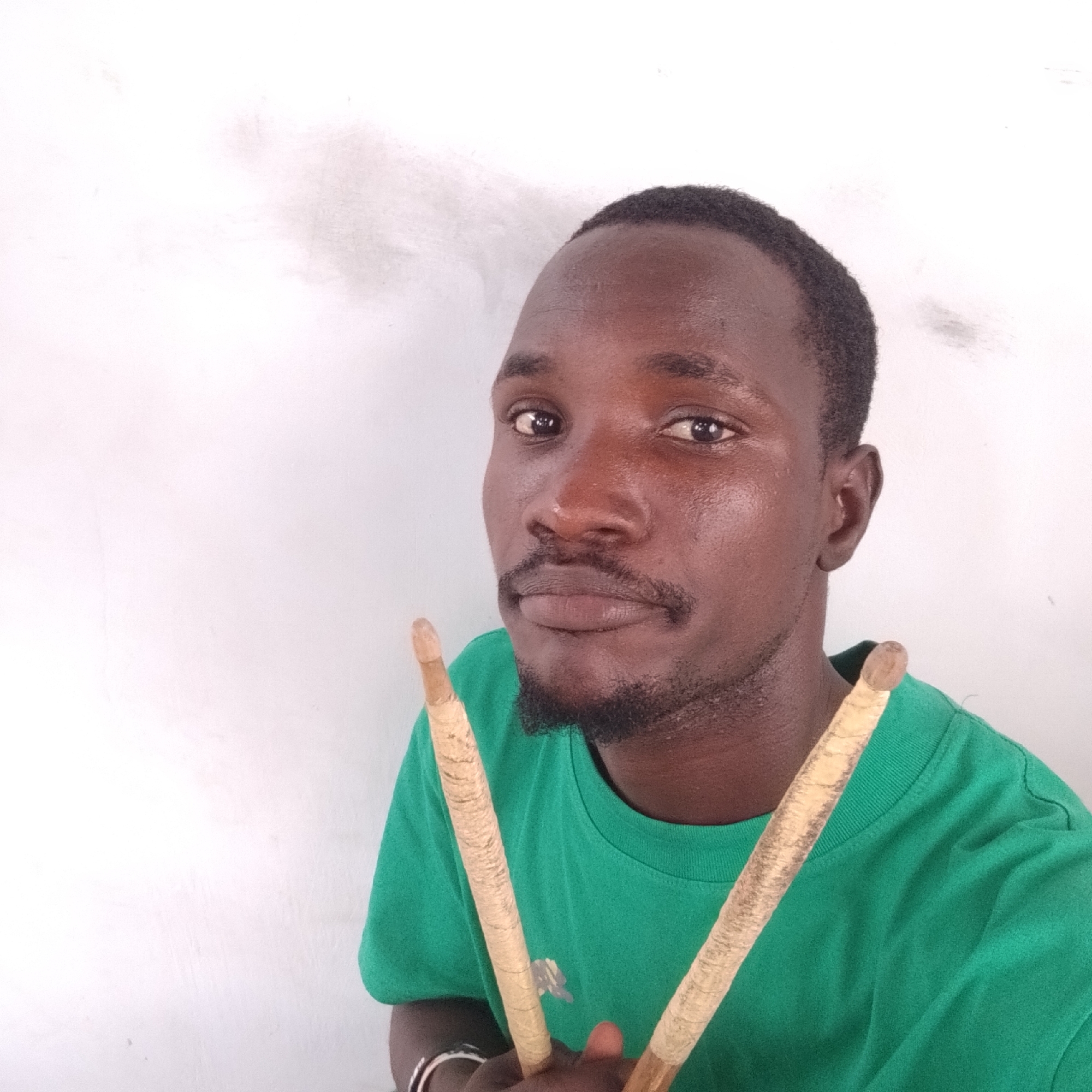 A man with dark skin, a mustache, and a goatee stands in front of a white, textured wall. He is wearing a green t-shirt and holding two drumsticks. The drumsticks are light brown with gold wrapping around the handles. The man is looking directly at the camera. The background appears to be an outdoor wall in Iranla, Nigeria.