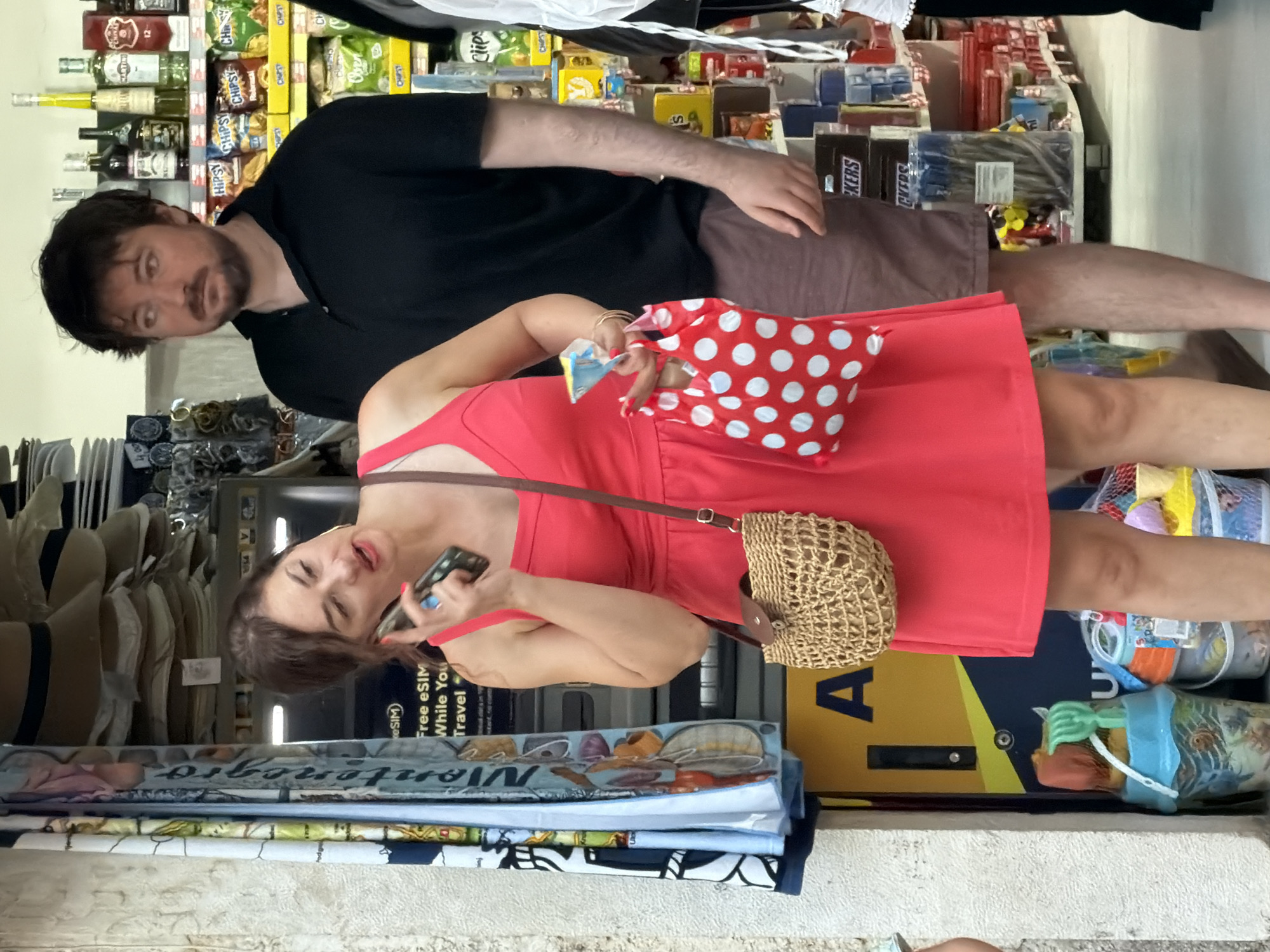 A woman in a bright red dress is standing on a street in Budva, Montenegro, holding a cell phone to her ear and a small red and white polka dot bag in her right hand. She also has a woven purse with a brown strap on her left shoulder. Beside her, a man in a black polo shirt and brown shorts stands with his arms down. In the background, a store shelf is stocked with various items, including bottles of alcohol and bags of chips. To the left of the woman, a yellow kiosk-like structure with the letter "A" on it is visible, as well as several buckets filled with children's beach toys. The scene appears to be a casual outdoor shopping area.