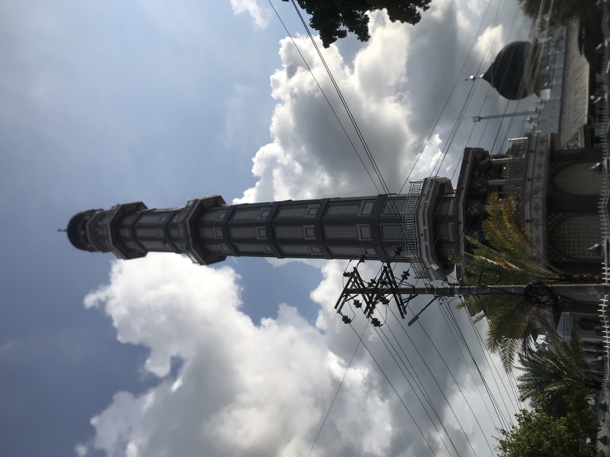 The image shows the Baiturrahman Grand Mosque Minaret in Keudekruenggeukueh, Indonesia. The minaret, a tall, grey, multi-tiered structure with a dome at the top, is the central subject. Power lines and a utility pole are visible in the foreground, intersecting the view of the minaret. The sky is partly cloudy. Parts of the mosque are visible on the right, including a dome. Trees are present in the foreground and background.