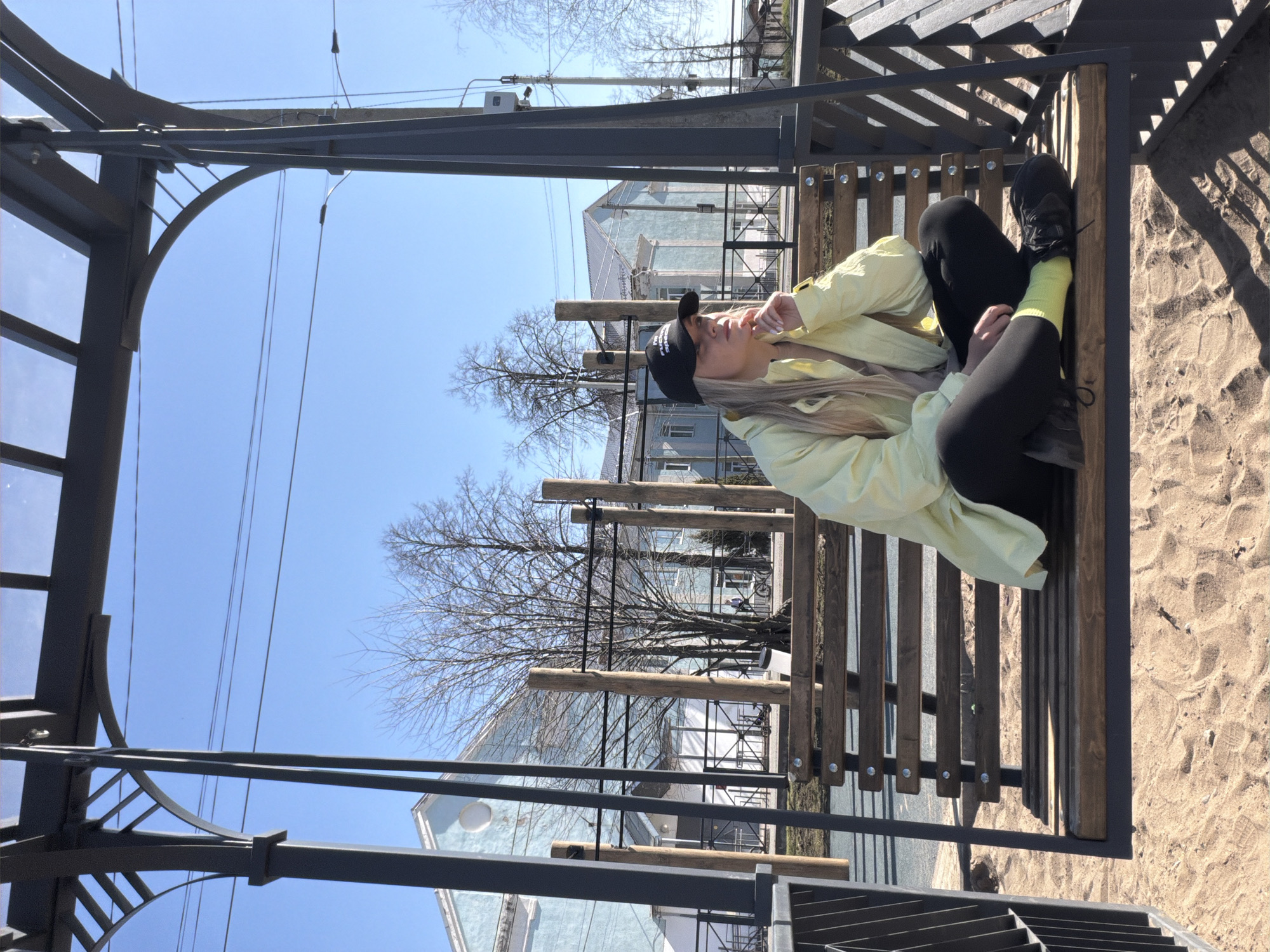 A woman with long, light-colored hair wearing a black baseball cap, a light yellow jacket, black pants, and black shoes sits cross-legged on a wooden bench within a metal structure. The structure has a roof and side walls, and is situated in a park or recreation area with a sandy surface. Behind the structure, trees with bare branches are visible, and a light blue building with a gray roof stands in the background. Power lines run across the sky. The image is taken in daylight. The location is most likely the city of Votkinsk, Russia, based on the user request.