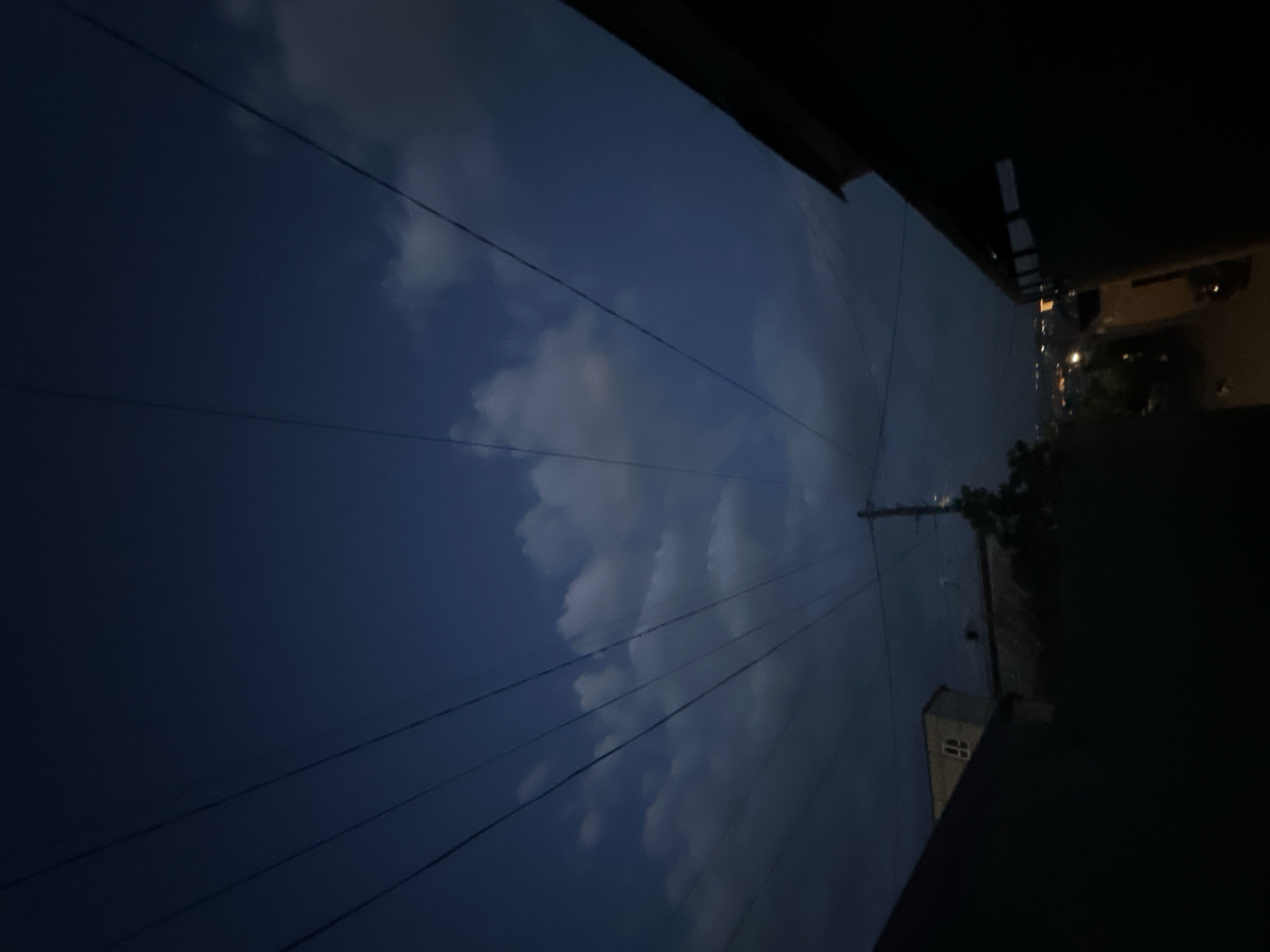The image is taken from a low angle, looking up at the sky. Power lines crisscross the frame against a backdrop of a dark blue evening sky with scattered white and grey clouds. On the right side, the edge of a building and a street are visible, both shrouded in darkness. The dim lights of buildings can be seen further down the street. The image suggests an urban environment, likely taken in the city of Yerevan, Armenia, based on the architectural style and street layout. There are no people visible.