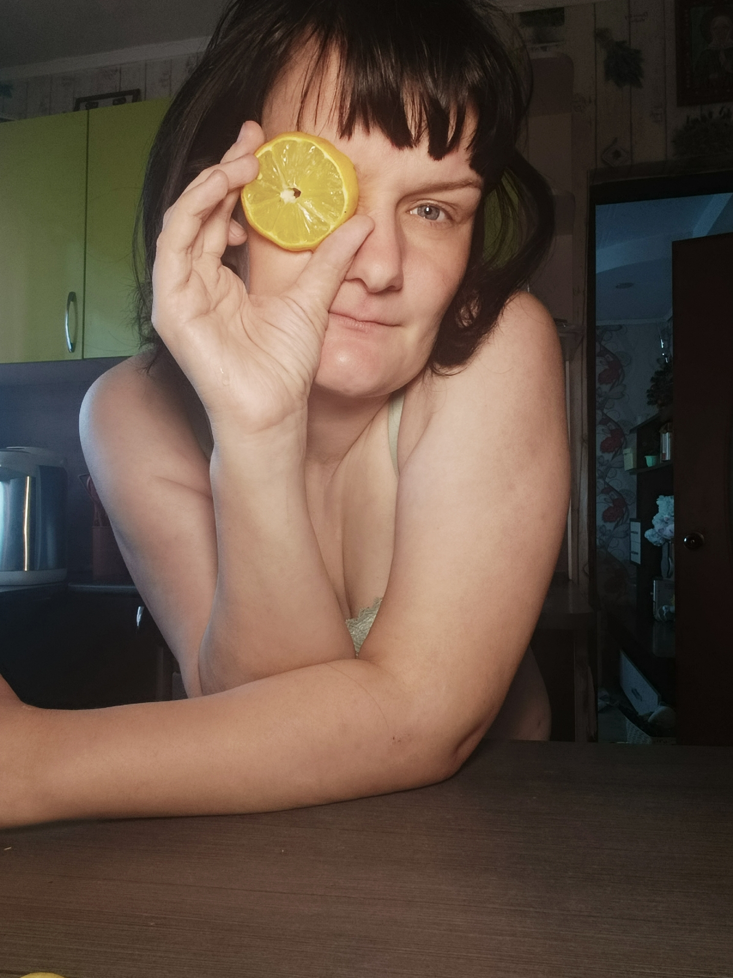A woman with dark hair and bangs partially covers her left eye with a slice of lemon. She is indoors, leaning on a wooden table. In the background are kitchen cabinets, a kettle, and a partially open doorway leading to another room with wallpaper. The location is Фоки, Russia.