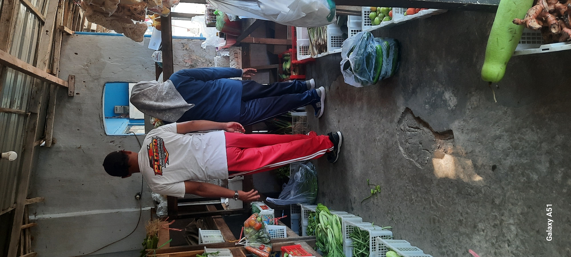 The image shows a market stall with two people, a man and a woman, standing. The man, wearing a white t-shirt with a logo and red pants, is facing away from the camera and towards the right. The woman, wearing a blue long-sleeved shirt and a grey headscarf, is also facing away from the camera, to the right of the man. The stall is filled with various fresh produce, including vegetables like leafy greens, cucumbers, and what appear to be gourds, displayed in white plastic crates and bags. The floor is concrete, with visible cracks and a distinct sunlight pattern cast on it. The stall structure is made of wood and corrugated metal. In the background, a doorway leads to a room with blue walls and some electrical equipment visible. The overall impression is a typical Indonesian market setting. The text "Galaxy A51" is visible in the bottom left corner of the image.