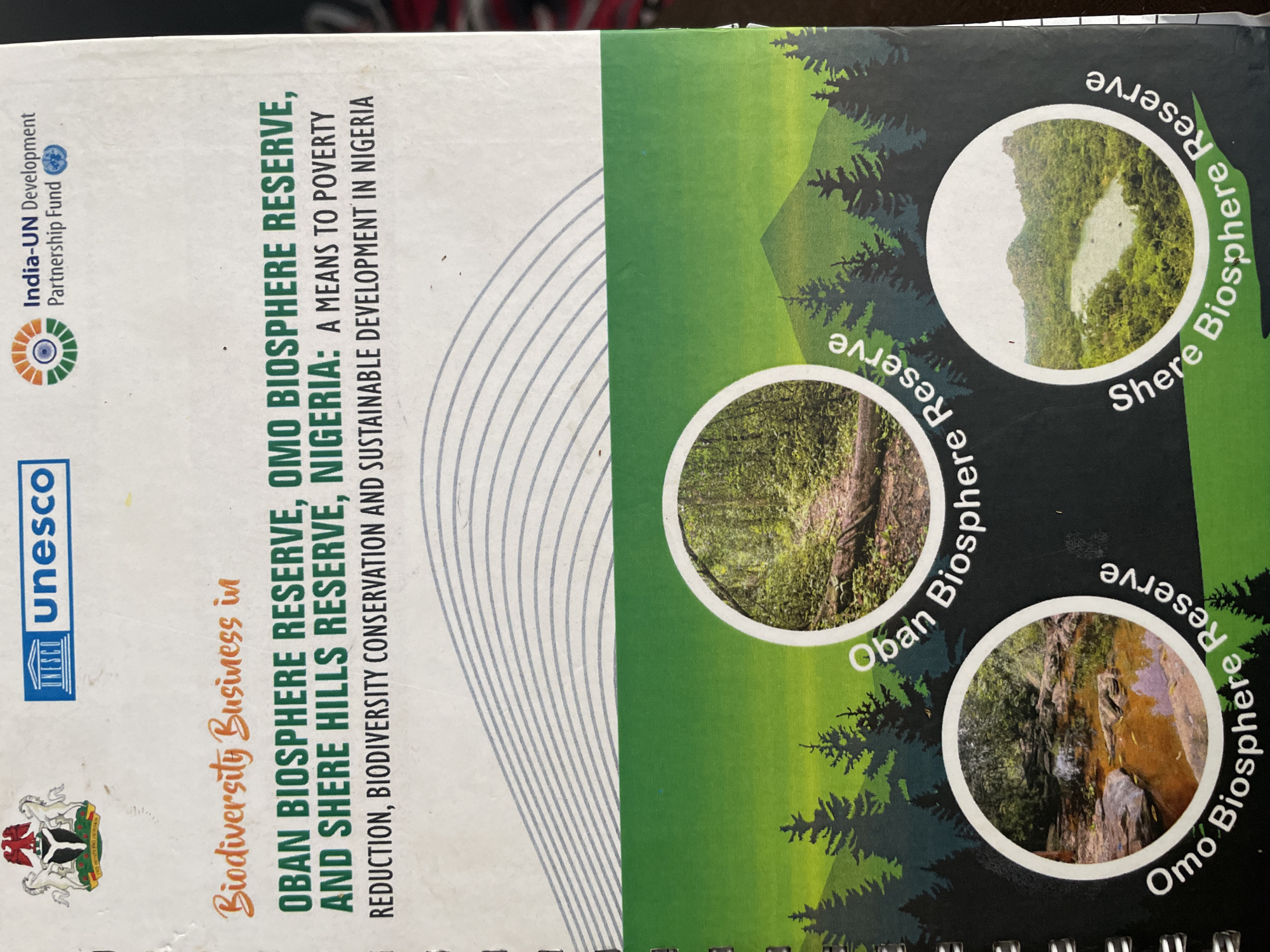 The image is a book cover or brochure that discusses biodiversity in Nigeria. The cover features the Nigerian coat of arms, the UNESCO logo, and the India-UN Development Partnership Fund logo. The title reads "Biodiversity Business in OBAN BIOSPHERE RESERVE, OMO BIOSPHERE RESERVE, AND SHERE HILLS RESERVE, NIGERIA: A MEANS TO POVERTY REDUCTION, BIODIVERSITY CONSERVATION AND SUSTAINABLE DEVELOPMENT IN NIGERIA." The lower portion of the image contains three circular photographs showcasing natural landscapes, each labeled with the name of a reserve. These photographs depict a forest path with a fallen log, a rocky stream with orange-tinted water, and a panoramic view of a lake surrounded by lush green hills. The overall design is green and white, with stylized trees framing the circular images. There is no visual information within the image that places any part of it specifically in the city of Jos, Nigeria.
