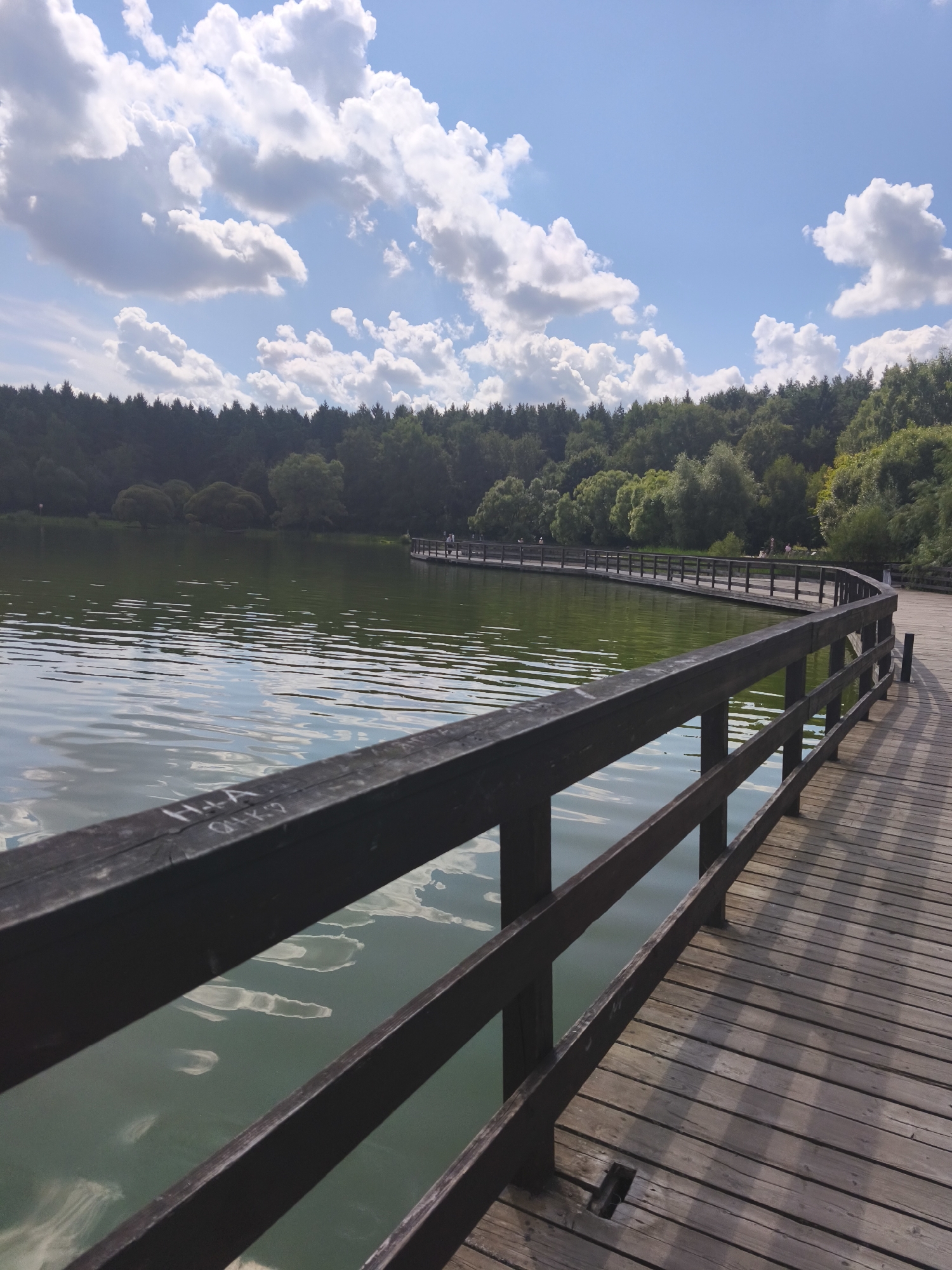 A wooden boardwalk curves along the edge of a tranquil lake, with a dense forest forming the backdrop. The water is a placid green, reflecting the scattered white clouds against a bright blue sky. The boardwalk is made of weathered planks and features dark wooden railings. In the distance, on the boardwalk, a few individuals are visible, appearing to be strolling. The scene conveys a sense of peaceful nature within an urban setting. This appears to be the Serebryany Bor park in Moscow, Russia, a popular recreational area featuring walking paths around its lakes and rivers.