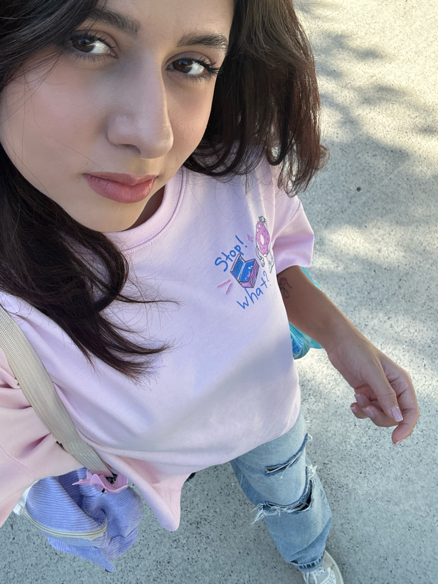A medium shot captures a young woman from the chest up, holding a purple and blue bag over her shoulder. She wears a light pink t-shirt with a cartoon graphic of a donut and a laptop, reading "Stop! What?". Her blue jeans have distressed rips. Her left arm is visible, and a subtle tattoo can be seen on her forearm. Her dark hair frames her face, and she has subtle makeup on. The background is an outdoor, paved area with dappled sunlight creating shadows. The scene appears to be a casual outing, possibly in the Sabail Raion district of Baku, Azerbaijan, given the pavement and lighting conditions often found in urban outdoor spaces there.