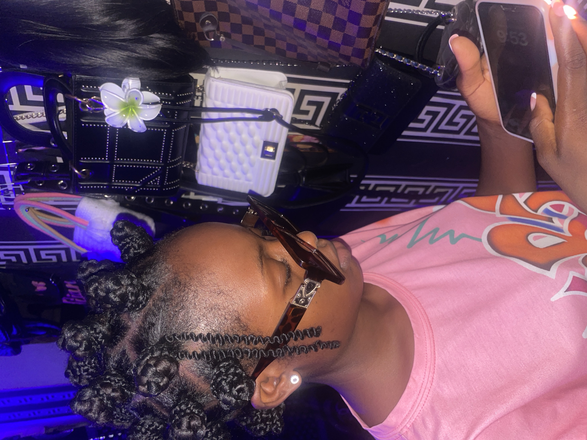 A person wearing sunglasses and a pink t-shirt is holding a smartphone in Lokoja, Nigeria. The phone displays the time as 9:53. Several handbags, including a black studded purse with a white flower and a checkerboard bag, are arranged on a patterned surface. The person has braided hair styled into knots.