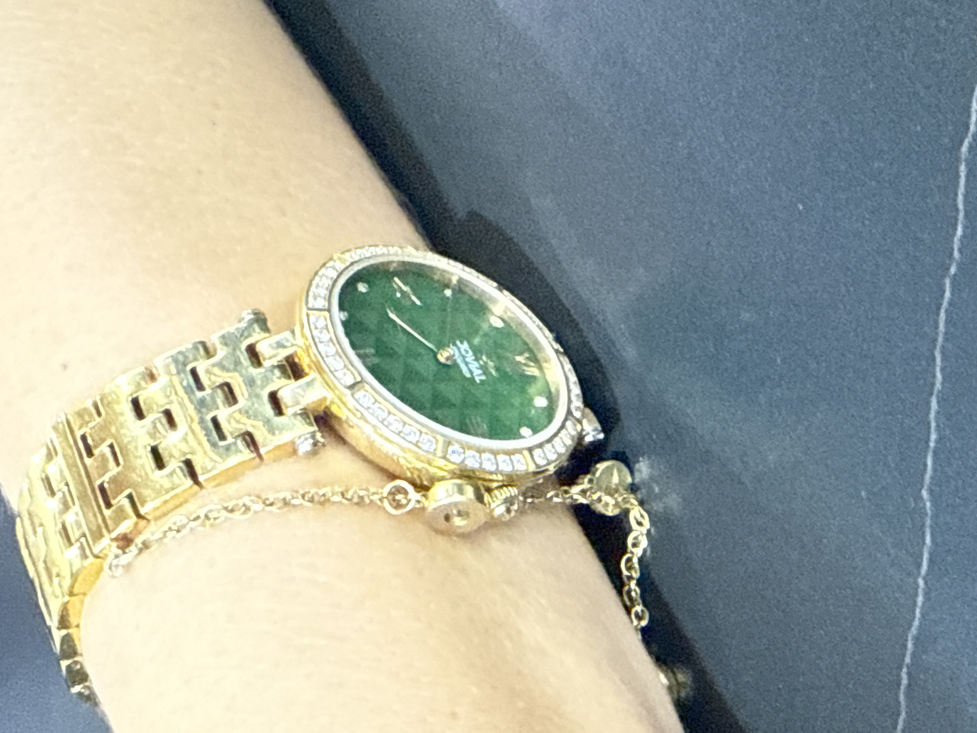 A close-up, eye-level shot shows a gold watch with a green face and diamond accents on a person's wrist. The watch band is made of interlocking gold links, and a delicate gold chain extends from the band to a small charm. The background is a dark, textured surface, possibly a table. The watch is positioned on the wrist in a way that suggests it is being worn. The image does not provide any information about the location of Dubai, United Arab Emirates.