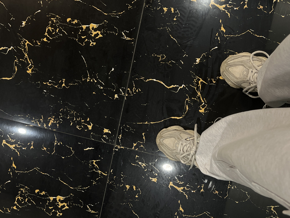 The image depicts an overhead view of a person's lower legs and feet positioned on a glossy, black tiled floor with intricate gold veining, resembling black Portoro marble. The floor is comprised of large, square or rectangular tiles with dark grout lines, reflecting a bright, circular light source from above near the upper left quadrant. The individual is wearing light grey, relaxed-fit trousers or sweatpants. On their feet are light beige or off-white athletic sneakers with a chunky sole and mesh upper, featuring visible New Balance branding. The shoelaces are tied. The person is in a static standing posture. The luxurious nature of the flooring suggests an indoor environment such as a high-end hotel lobby, a modern upscale retail establishment, or a residential interior within Nha Trang, Vietnam.