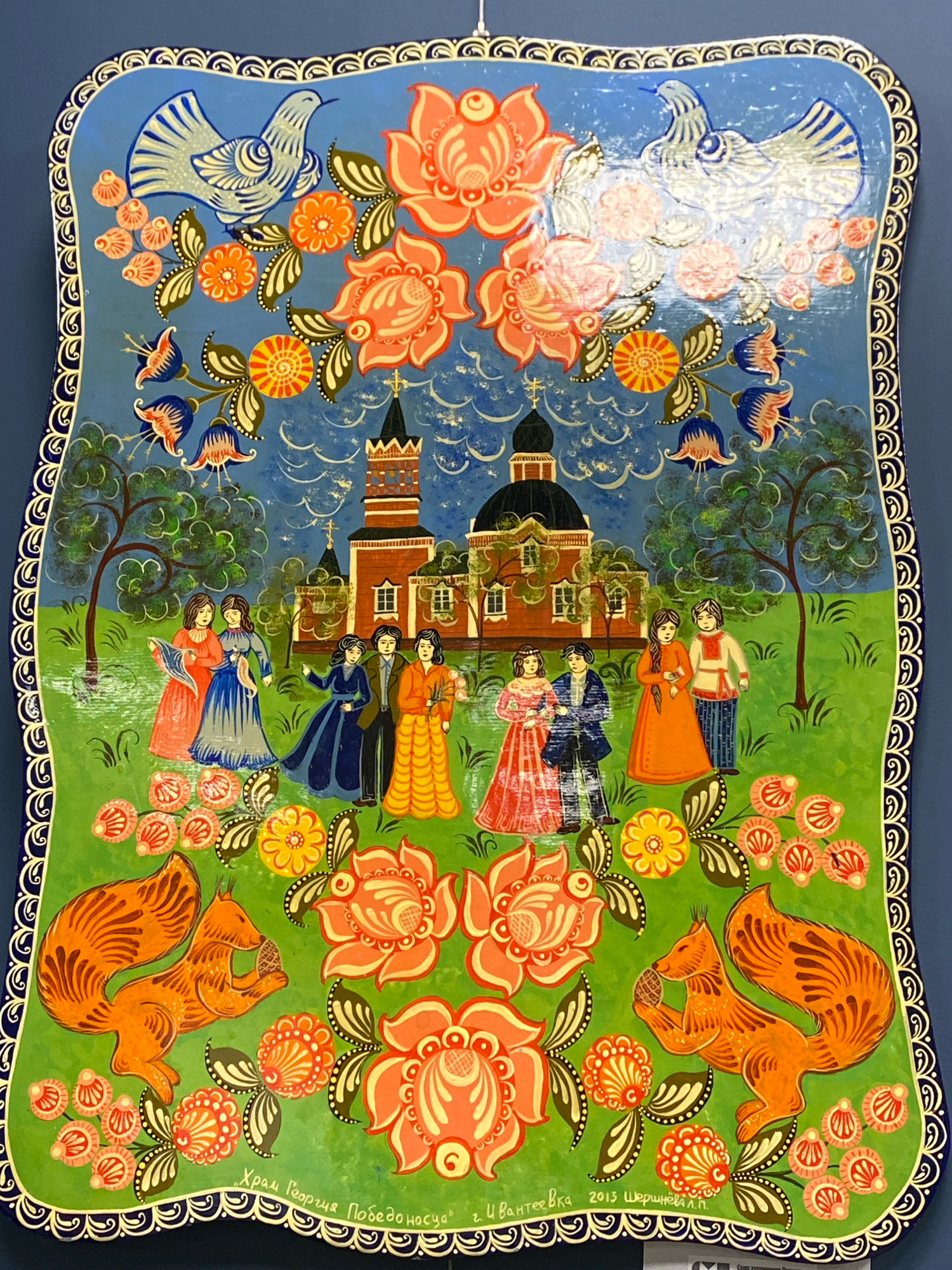 This is a brightly colored, folk art painting on a wooden surface depicting a scene set in a green, grassy area with a blue, cloudy sky. In the background, a red brick building with a dark dome, resembling a church, stands prominently. Flanking the church are two trees with green foliage. In front of the church, a group of seven figures, dressed in traditional clothing, are gathered. The women wear long, flowing dresses in various colors, including red, blue, and orange, while the men are depicted in shirts and pants. Two of the women hold fabric or scarves.
The foreground is adorned with vibrant floral motifs in shades of orange, pink, yellow, and blue, interspersed with green leaves. Two orange squirrels, with bushy tails, are a central feature in the lower portion of the painting, each holding a brown nut or cone.
Along the bottom edge of the painting, there is Russian text, which, based on the visible characters, includes "г. Ивантеевка" (g. Ivanteyevka), indicating that the scene or the artwork is associated with the city of Ivanteyevka, Russia. Further text, partially obscured, appears to be a date, likely "2013". The entire painting is framed by a decorative border with swirling patterns in blue and white. The painting appears to be a decorative plaque or tray.