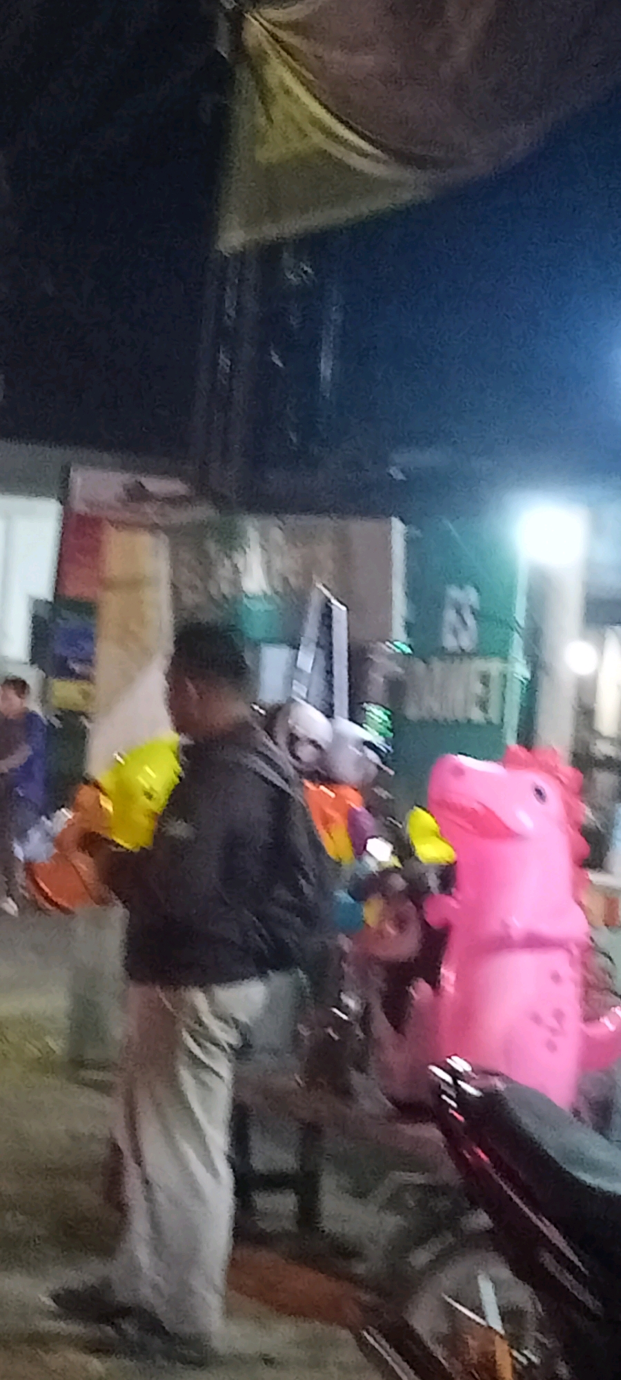The image shows an outdoor scene at night, possibly a street or marketplace, in Singogalih, Indonesia. In the foreground, a person wearing a dark jacket and light-colored pants is holding a bright yellow inflatable toy. To the right of this person, there is a large pink inflatable dinosaur toy. Behind them, other inflatable toys in various colors, including orange, purple, and blue, are visible, some attached to what appear to be motorcycles. In the background, there are signs and structures, including a green sign with white lettering that partially reads "MARKET". The overall lighting suggests it is nighttime with artificial light sources present.