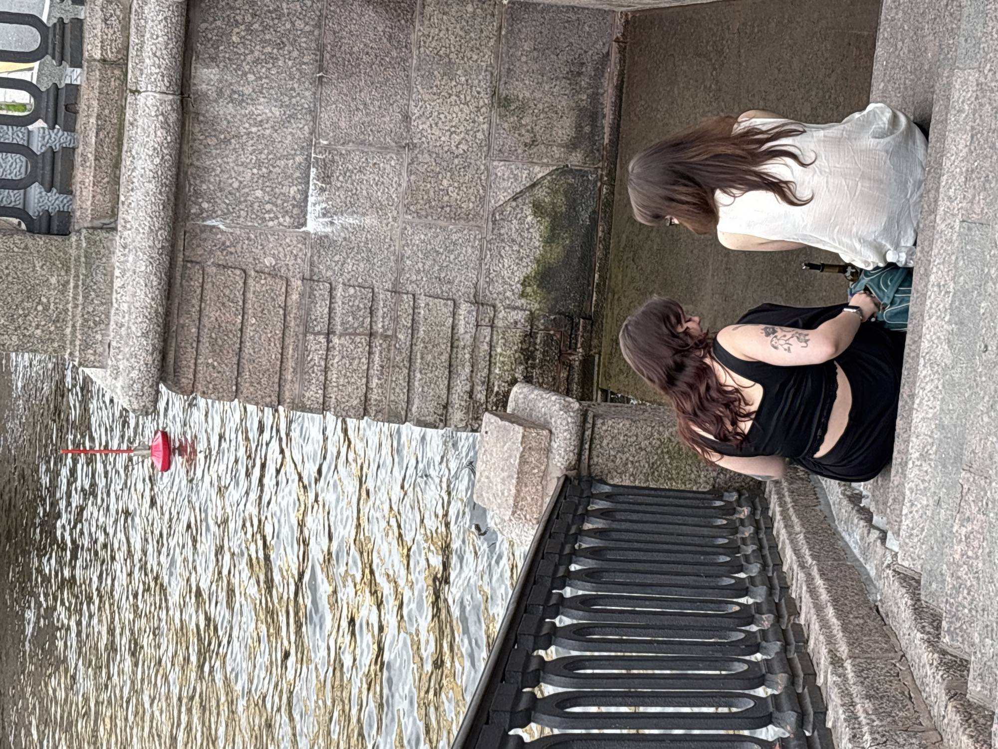 An elevated view captures two women by a granite embankment, overlooking a body of water, likely a canal or river in Saint Petersburg, Russia. The image is oriented sideways, with the water on the left and the embankment on the right.
In the foreground, a woman with long, dark brown hair wears a black top, revealing a tattoo on her right shoulder/arm and a watch on her left wrist. She is seated or kneeling on the stone steps, looking downwards towards the water. Behind her, another woman with long, lighter brown hair is also seated or kneeling, wearing a white top. She has a dark green object, possibly a bottle or bag, next to her. Both women have their backs partially to the viewer, appearing to observe something in the water or simply relaxing by the embankment.
The setting consists of rough-hewn, light grey granite blocks forming steps that descend towards the water and a wall along the right. A dark, ornate metal railing runs along the left edge of the embankment, separating it from the water. The water itself is rippled, reflecting sunlight with shimmering patterns, and a red-tipped buoy or marker floats in the middle-left of the frame. The scene suggests a calm, daytime atmosphere. No visible text is present.