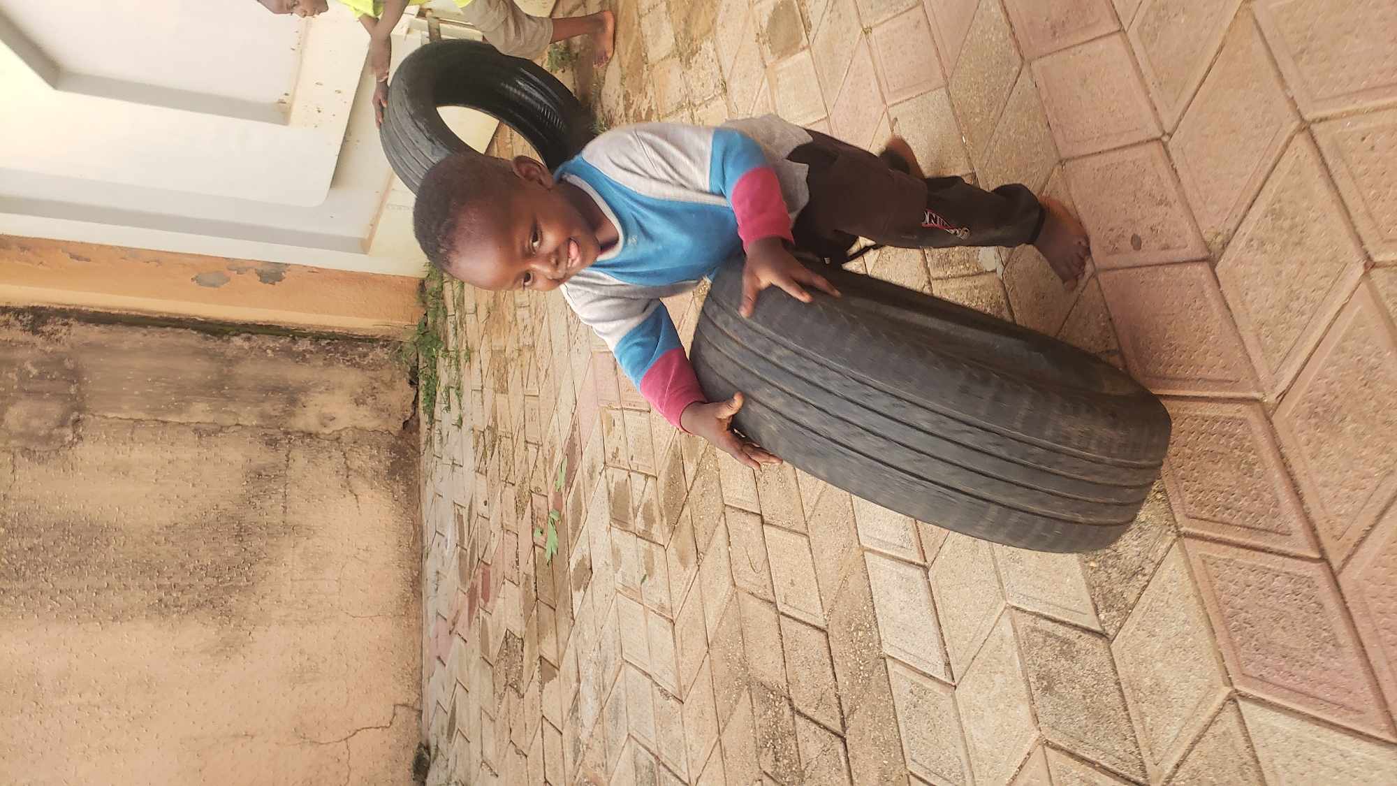 A young Black child is pushing a large, worn tire across a paved courtyard in Sabon Tasha, Nigeria. The child, with short dark hair and a playful expression, is wearing a gray long-sleeved shirt with blue and pink accents on the sleeves, and dark pants. Their tongue is slightly sticking out, suggesting effort or excitement. Their hands are gripping the rubber tire, which shows significant tread wear. The courtyard is paved with light brown and pink diamond-shaped tiles, some of which appear to be slightly uneven. A second child, wearing a yellow shirt and brown shorts, is partially visible in the background, also near a tire. The environment features a textured, light beige wall with visible cracks and some dark staining, possibly due to age or weathering. To the right, a white structure with a curved edge is visible, suggesting part of a building or play area. Sparse green vegetation is growing between some of the paving stones. The scene is outdoors, with natural lighting.