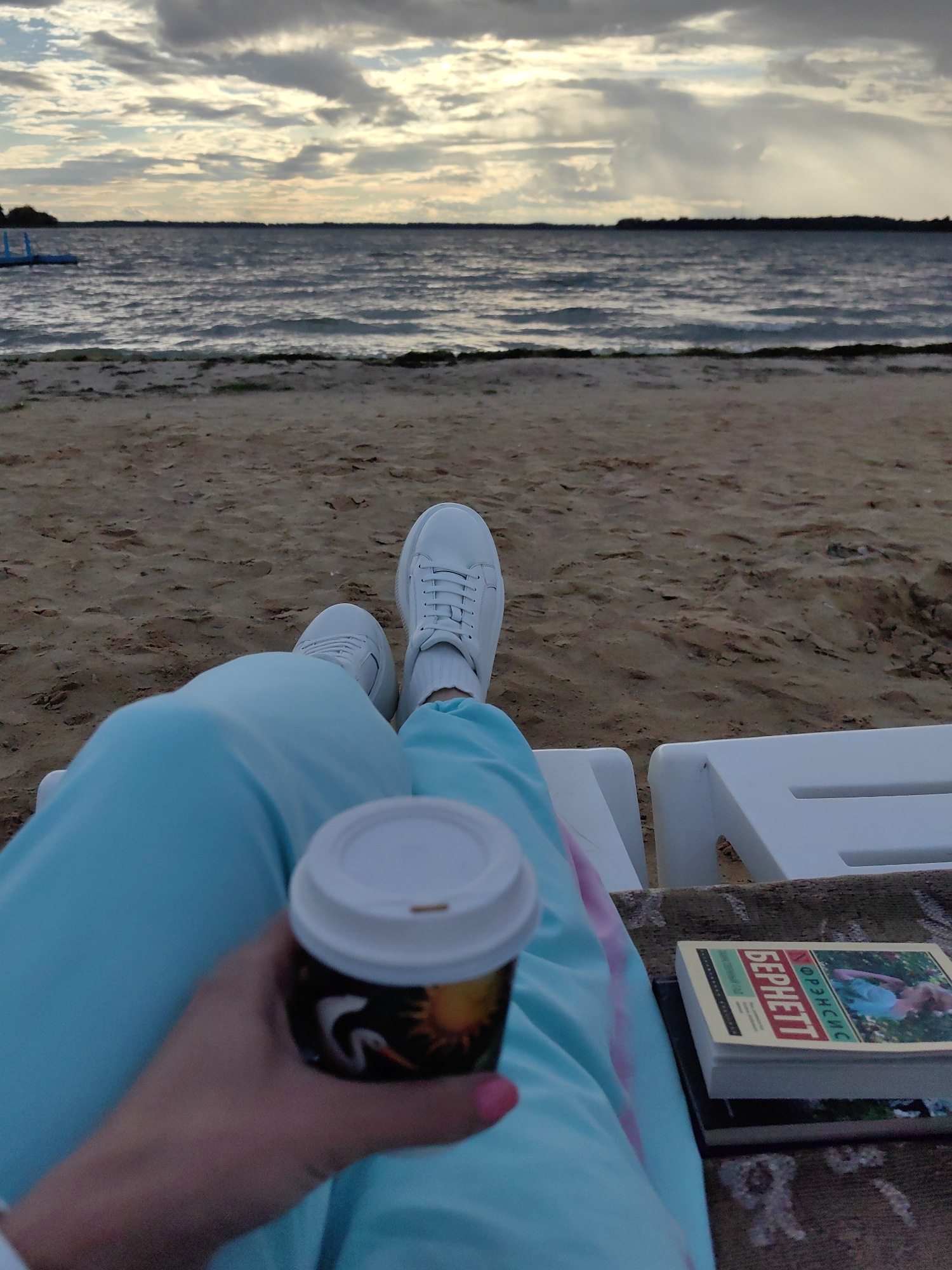 A person is sitting on a white lounge chair on a sandy beach, facing a body of water. They are wearing light blue sweatpants and white sneakers. In their right hand, they hold a disposable coffee cup with a white lid and a graphic design on the side. To their right, on a patterned rug, are two books. The larger book, on top, has a colorful cover with text in Russian and an image of a person. The body of water is choppy with waves, and the sky is overcast with dramatic clouds. In the distance, a dark, tree-lined shore is visible, and to the left, a blue structure, possibly a dock or water feature, protrudes into the water. The overall impression is of a person enjoying a quiet moment by the water. The location is identified as Myadzyel, Belarus.