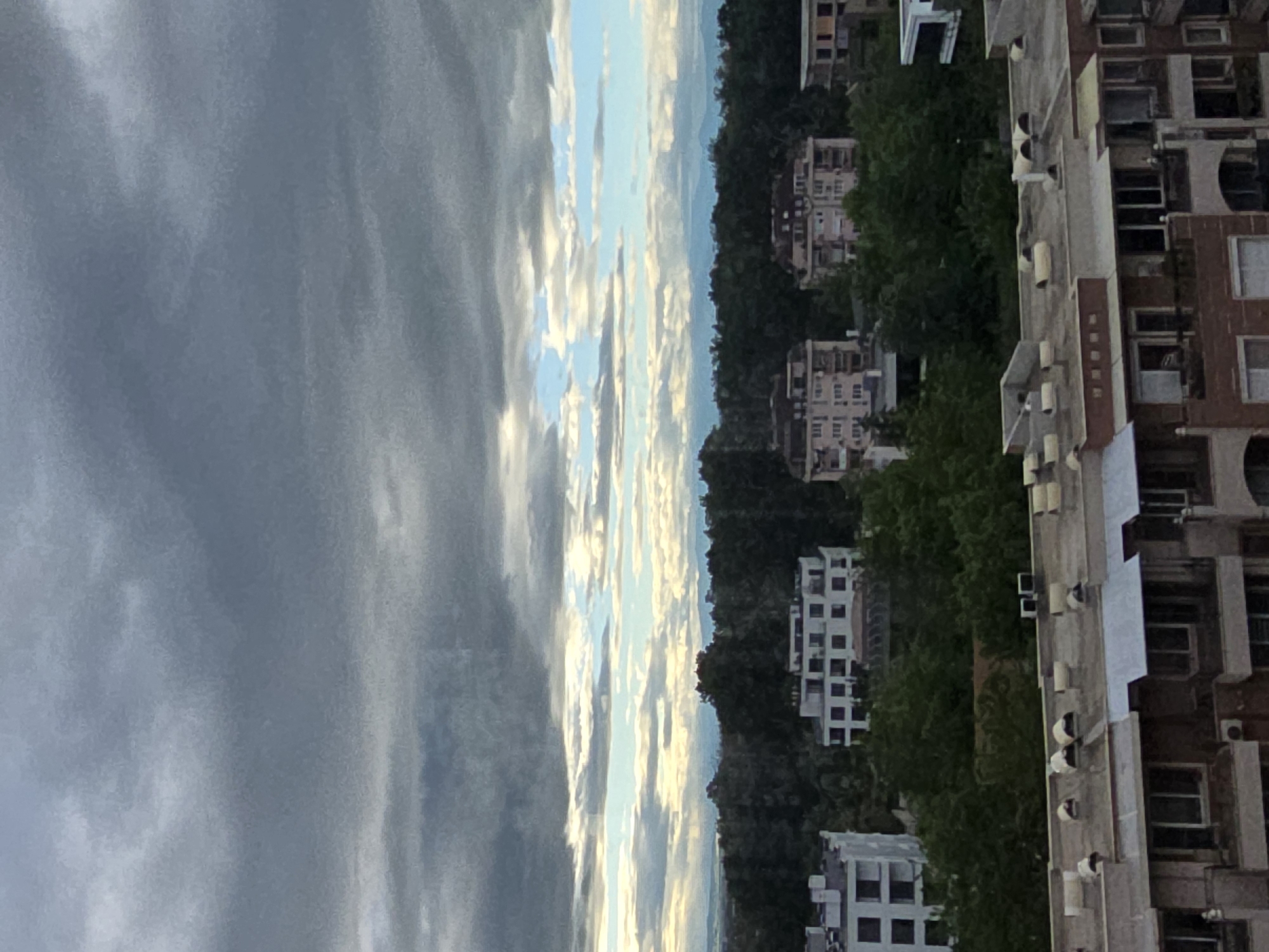 The image shows a cityscape under a cloudy sky. On the right side of the frame, a tall building with multiple floors and windows is visible. Further away, amidst green trees, are several other buildings of varying sizes. The sky occupies a significant portion of the image, filled with layered clouds and hints of blue. The scene appears to be set in a dense urban area, although the specific location is unknown.