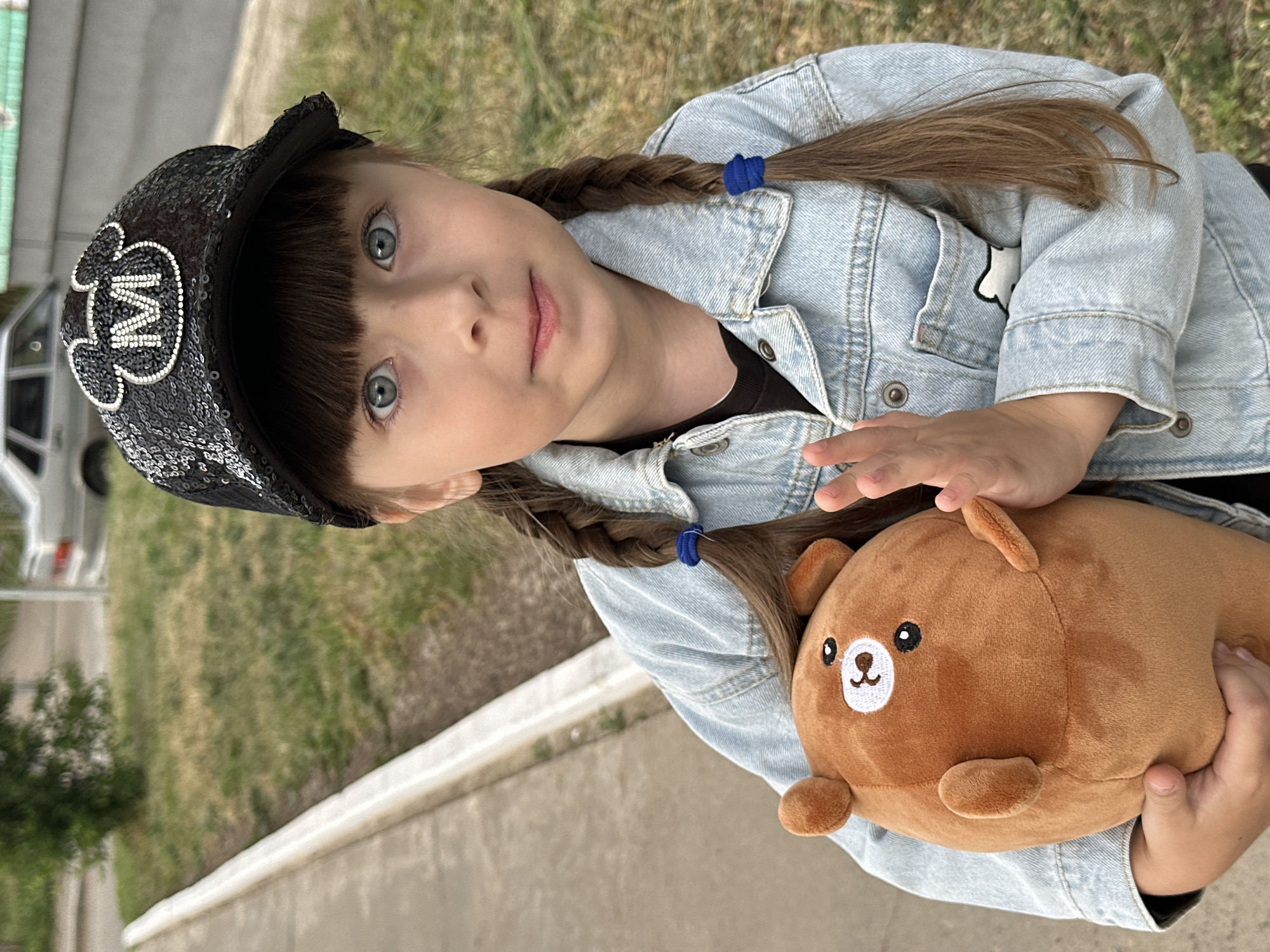 A young girl with light brown braided hair and striking green eyes is holding a stuffed brown bear. She is wearing a light denim jacket over a dark shirt and a black sequined baseball cap with a stylized "M" on it. The photograph appears to be taken outdoors in the city of Chaikovsky, Russia, based on the presence of typical urban features like a paved walkway, a low concrete wall or curb, grass, and a parked vehicle visible in the background. The girl looks towards the camera with a slightly inquisitive expression. She has her left hand raised slightly, almost touching the bear. The scene suggests a casual, everyday moment.