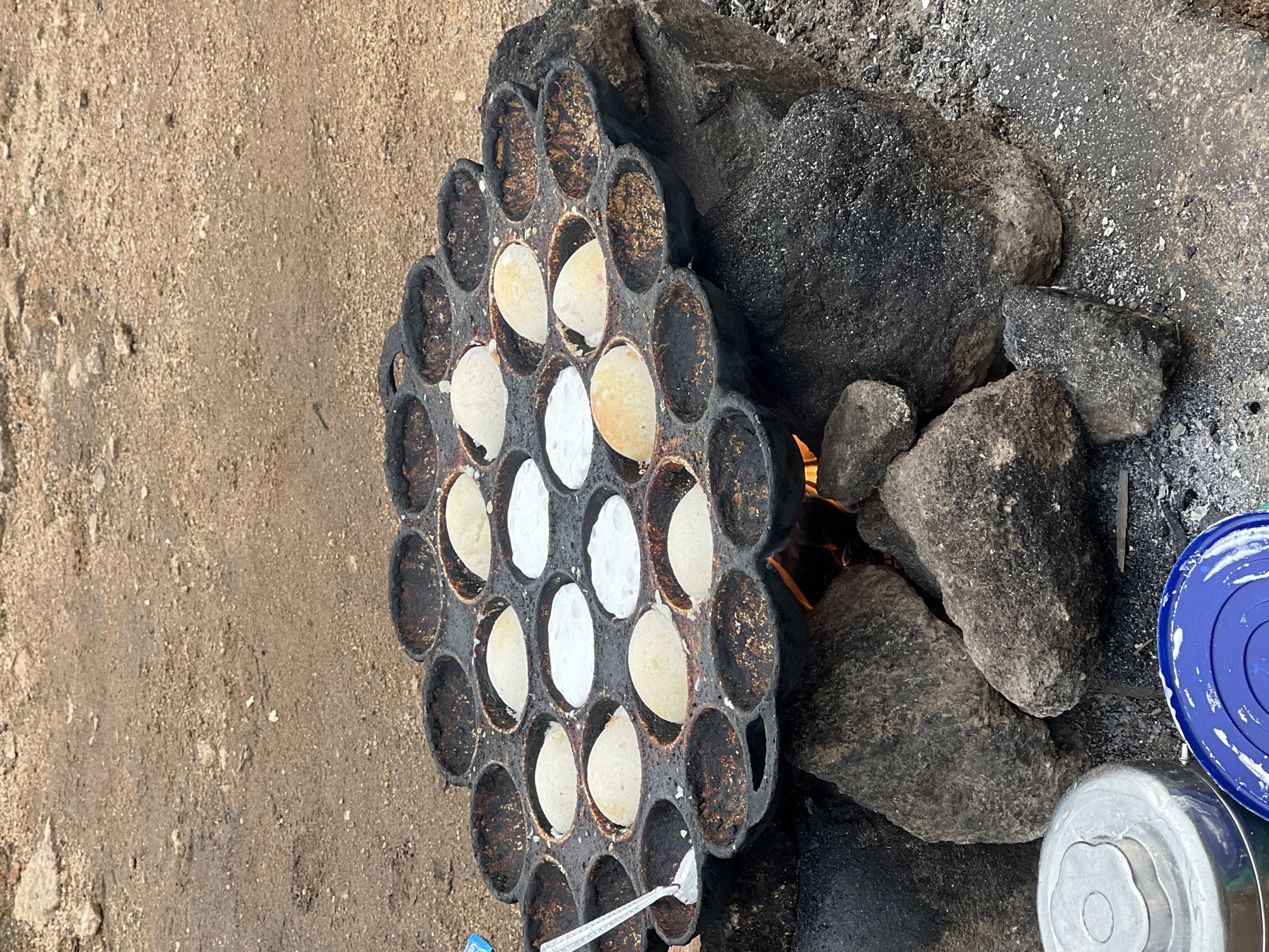 In the city of Jos, Nigeria, a round, black, cast-iron pan with numerous circular indentations is placed on a collection of dark gray rocks over a visible fire. The pan contains small, cooked food items resembling pancakes or fritters, with some appearing golden brown and others a lighter off-white color. Several indentations are empty, showing the dark, well-seasoned surface of the pan. A silver utensil with a long handle, possibly a spatula, rests in one of the indentations on the left side of the pan. The surrounding ground is composed of reddish-brown dirt and small pebbles. In the lower left corner of the image, a silver metal container with a patterned lid is partially visible, along with a blue plastic lid. The scene appears to be an outdoor cooking setup, likely for preparing traditional food.