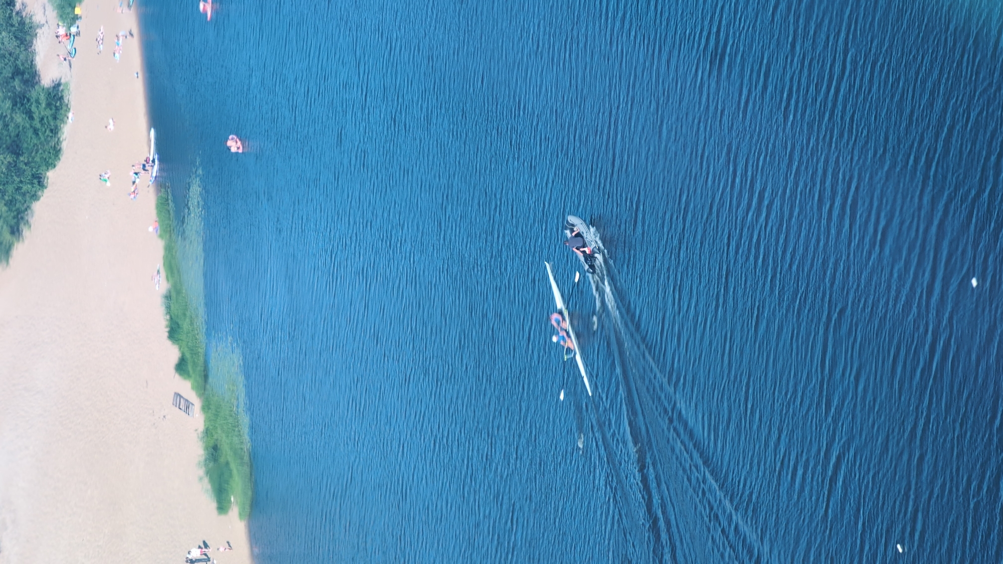 An aerial, top-down view shows a sunlit, sandy beach bordering a vast expanse of blue water. The beach is populated with numerous sunbathers, some lying on towels and others engaged in various beach activities. A line of green vegetation separates the sand from the water.
In the water, two individuals are participating in a watersport. One person is on a stand-up paddleboard near the shore, while another is in a sleek, white racing kayak, propelled forward by a person in the back. The kayak leaves a visible wake behind it, indicating movement. Further out in the water, a small, dark-colored object, possibly a boat, is also visible with a wake trailing behind it.
The scene suggests a pleasant day with clear skies, suitable for outdoor recreation. The bright sunlight casts clear shadows, and the water appears calm with gentle ripples. The location is identified as Nizhny Novgorod, Russia. No visible text is present in the image.