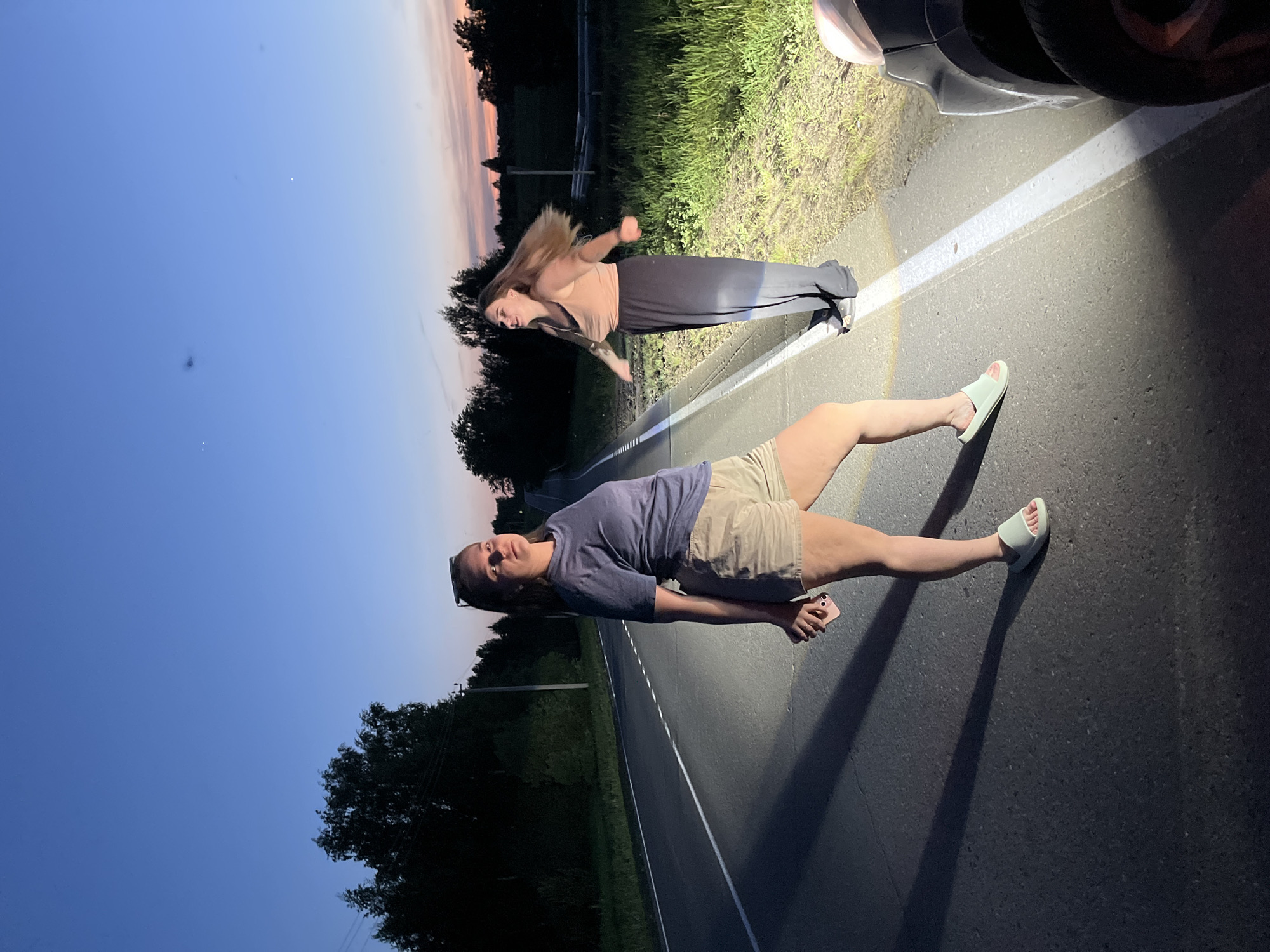 Two women are lying on a paved road at dusk. The first woman is wearing a navy t-shirt and tan shorts, and light-colored sandals. She is holding a pink object in her hands. The second woman is wearing a tan top and black skirt. The road is illuminated by the headlights of a car parked at the edge of the road. Green foliage is visible on either side of the road, and trees are visible in the distance. The sky is blue fading into peach color. The location is Ольховка, Russia.