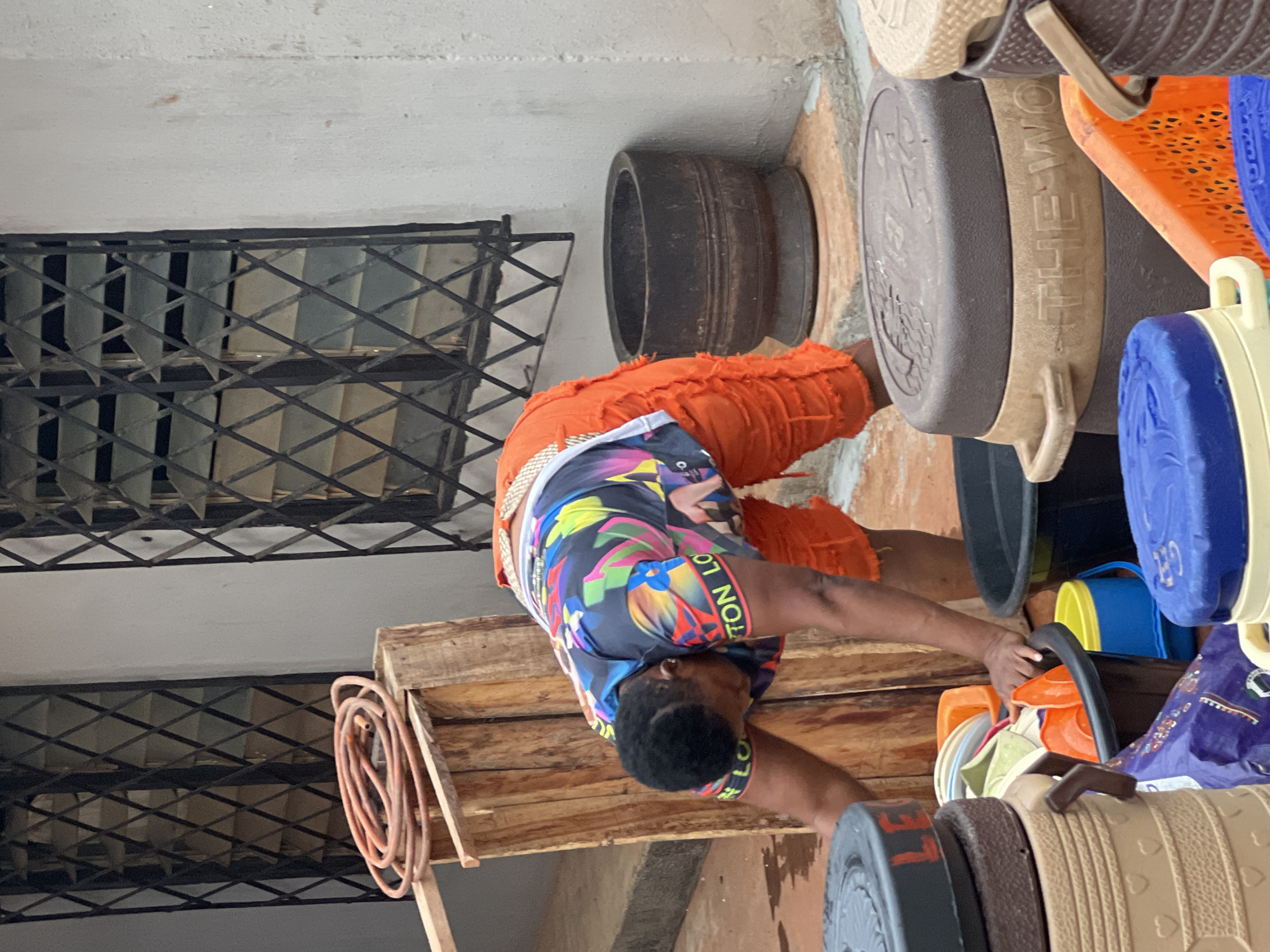 This image captures a vibrant market scene in Onitsha, Nigeria, focusing on a person interacting with a multitude of colorful plastic wares. The main subject is an individual, likely male, with short dark hair, wearing a brightly patterned t-shirt and ripped orange pants. The t-shirt features a colorful, abstract design, and partial text "LV.TION LO" is visible on the sleeve.
The person is positioned in a bent-over posture, appearing to be sorting, arranging, or selling a large collection of plastic buckets, tubs, and other containers. These items are stacked and displayed prominently around them, showcasing a diverse range of colors including blue, yellow, orange, beige, and various other hues. One beige container clearly shows the embossed text "THE WORLD". Another large, dark, barrel-like object is visible in the background, near the person's orange pants.
The setting appears to be a market stall or a semi-enclosed commercial space. Above and to the left of the person, a dark metal grate or ventilation system with a diamond pattern is visible against a light-colored wall or ceiling. A wooden pallet-like structure is also present, and a coiled orange hose can be seen resting on it. The overall impression is one of a busy marketplace specializing in household plastic goods. The lighting suggests it is daytime.