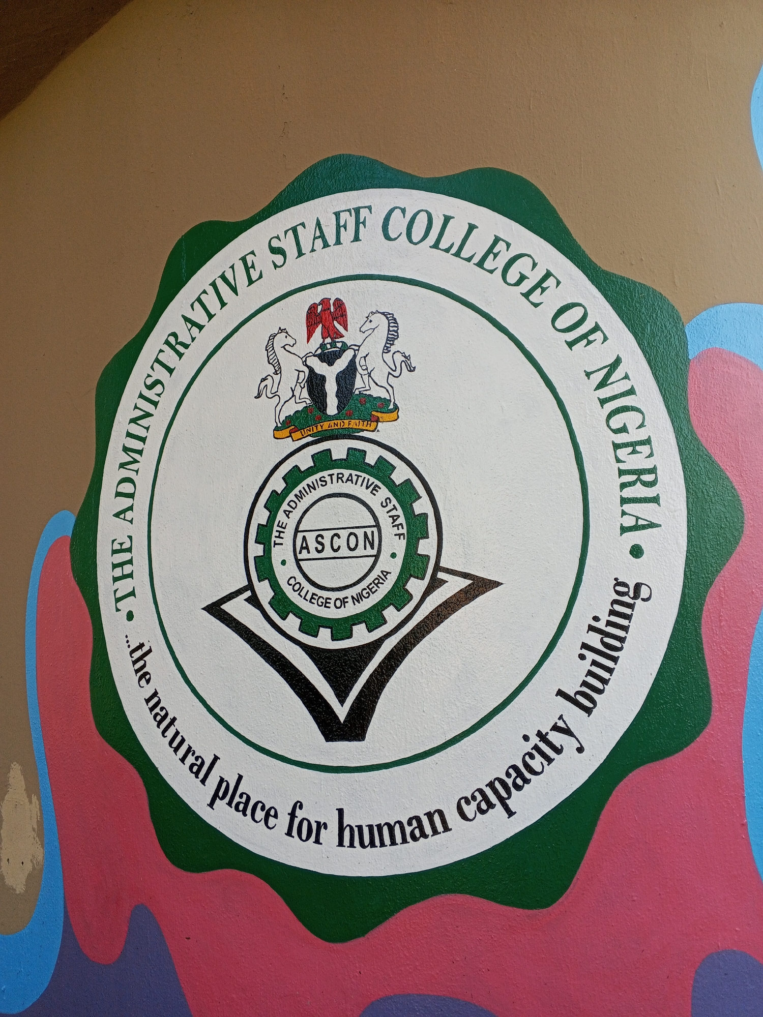 The image shows a painted emblem of the Administrative Staff College of Nigeria (ASCON). The emblem is circular with a green scalloped border. Within the border, text reads "THE ADMINISTRATIVE STAFF COLLEGE OF NIGERIA" at the top and "...the natural place for human capacity building" at the bottom. In the center of the circle is a representation of the Nigerian coat of arms above another smaller emblem. The inner emblem is a gear-like shape with the words "THE ADMINISTRATIVE STAFF" at the top and "COLLEGE OF NIGERIA" at the bottom, with "ASCON" in the center. Below this gear-like emblem is a black inverted chevron shape. The emblem is painted on a wall with a brown background and a section of colorful wavy patterns along the right side and bottom. The image does not include information about the specific city of Tawpo, Nigeria.