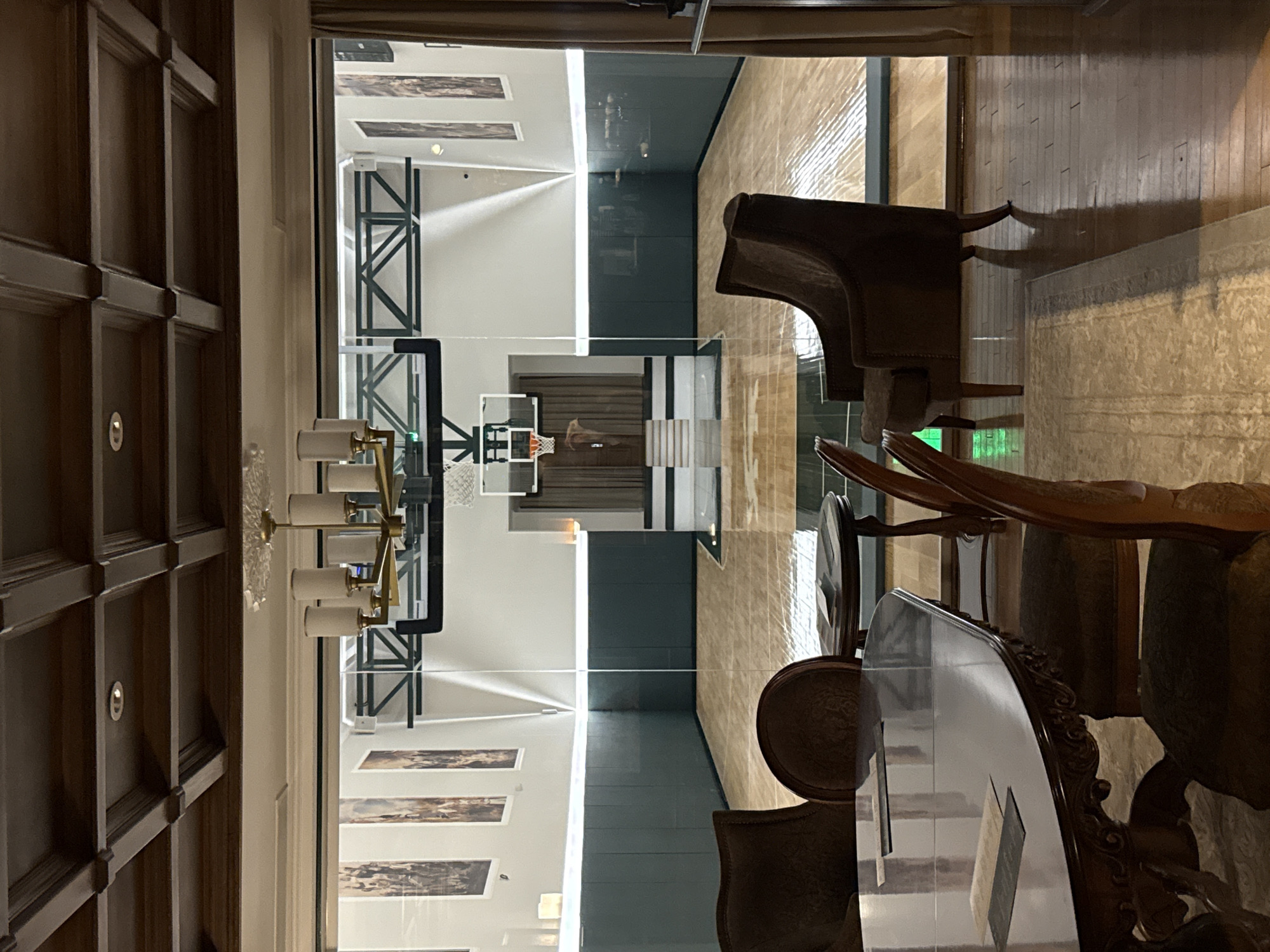 The image depicts an indoor basketball court viewed through a glass partition from a well-appointed sitting area. The court features a polished wooden floor, dark green wall padding, and a standard basketball hoop and backboard. The surrounding area is illuminated by overhead lighting and strip lighting along the walls. In the foreground, a dark wood dining table is set with papers, and several upholstered chairs are arranged around it, indicating a domestic or private recreational setting. The overall aesthetic suggests a luxurious and private space. The provided information states the location is Zhukovka, Russia, which implies this is likely a private residence or exclusive club.
