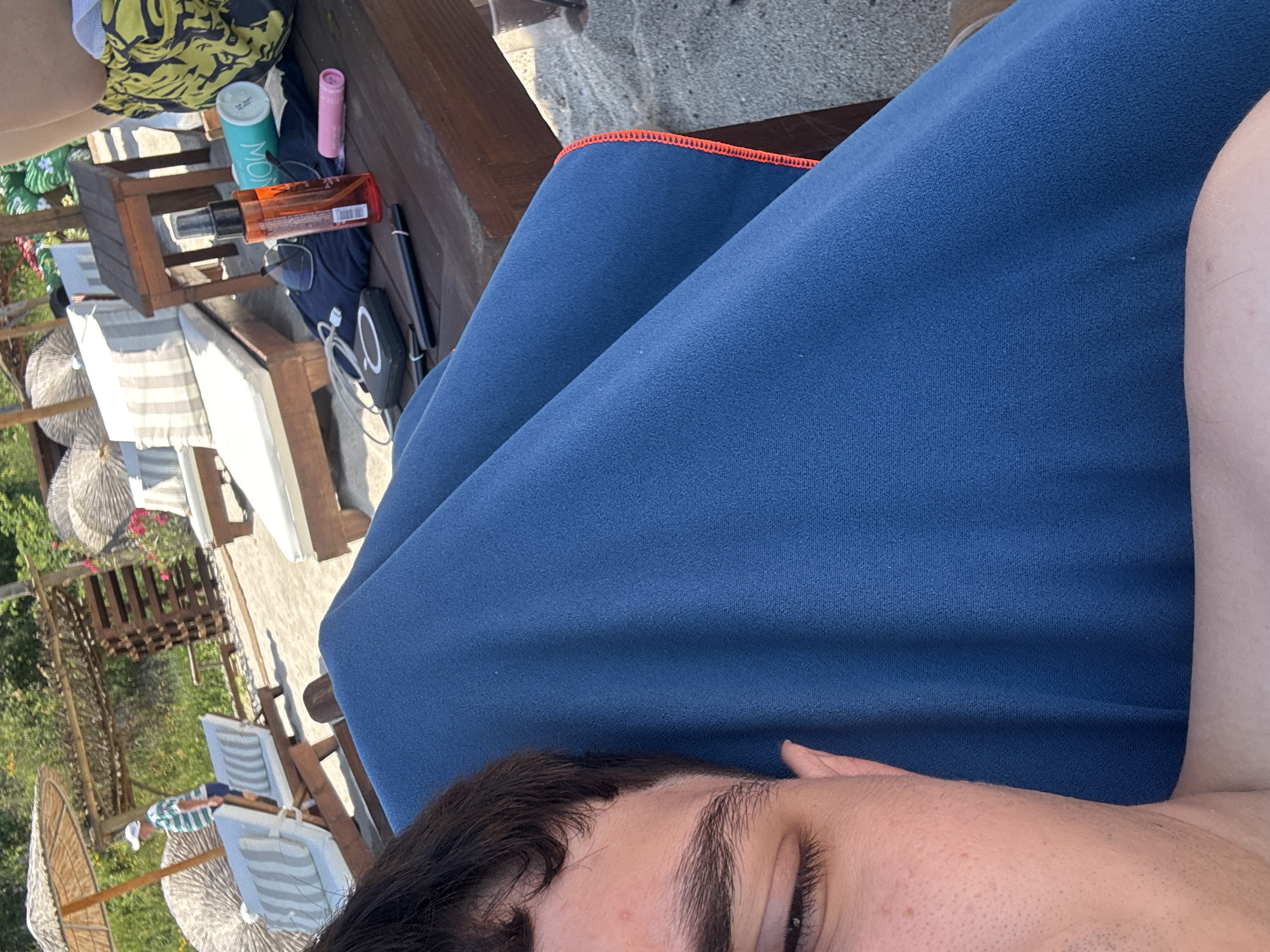 The image depicts a beachfront scene, likely in Varna, Bulgaria, characterized by sandy ground, lounge chairs, and umbrellas. In the foreground, a person's face and shoulder are visible, draped in a blue towel. On a wooden table in the midground, various objects are arranged, including sunglasses, a bottle of what appears to be sunscreen, a teal can with the word "MOK" on it, and a pink cylindrical item. Further back, a man in a striped shirt and white hat stands near a lounge chair. The environment suggests a recreational setting, possibly a beach resort.