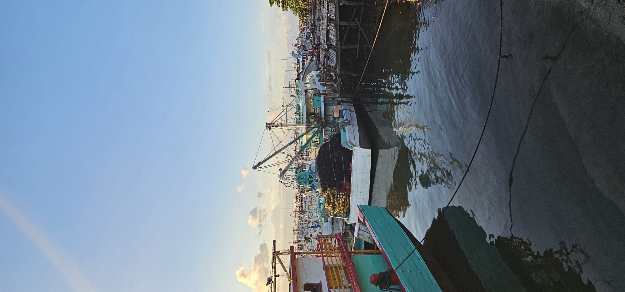 The image shows a marina in Denpasar, Indonesia. Numerous fishing boats are docked along the waterfront. Several of the boats have cranes. The water is dark, with reflections of the boats and structures. The sky is blue with a hint of orange on the horizon. A weathered wooden pier is visible near the upper edge of the frame.