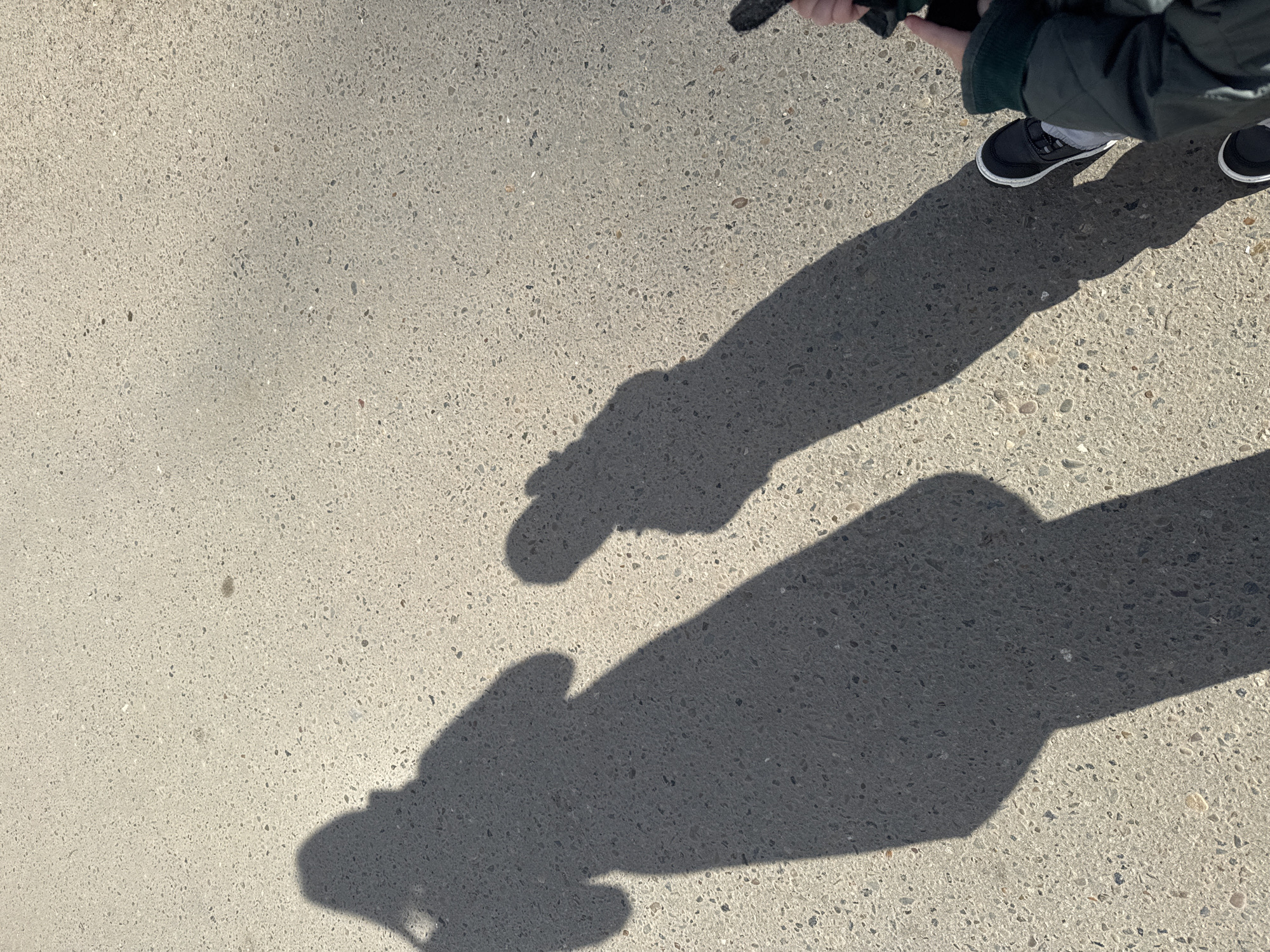 The image shows a concrete surface in full sunlight with the shadows of a person and an object. The person's lower legs, feet in black and white sneakers, and part of their green jacket are visible in the upper right corner. The shadows suggest the person is holding a small object in their hands. The exact location in Chaikovsky, Russia, cannot be determined from the image alone.