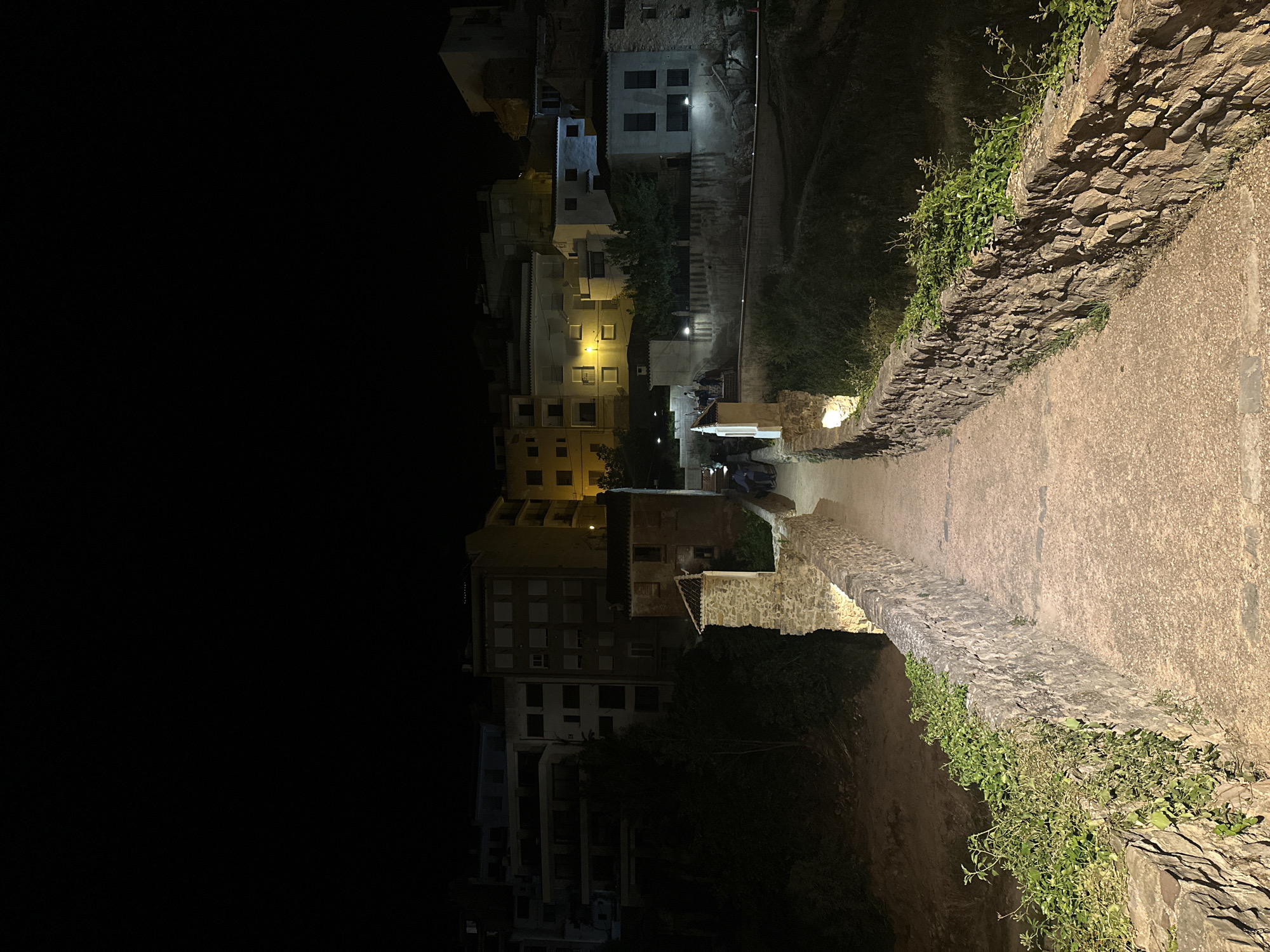 This is a nighttime image of a paved path leading away from a cluster of buildings in Montanejos, Spain. The path is bordered on both sides by stone walls covered in patches of green vegetation. The right side of the image is more brightly lit by ambient light. The buildings are illuminated by streetlights and light emanating from windows. The left side of the image is obscured by darkness.