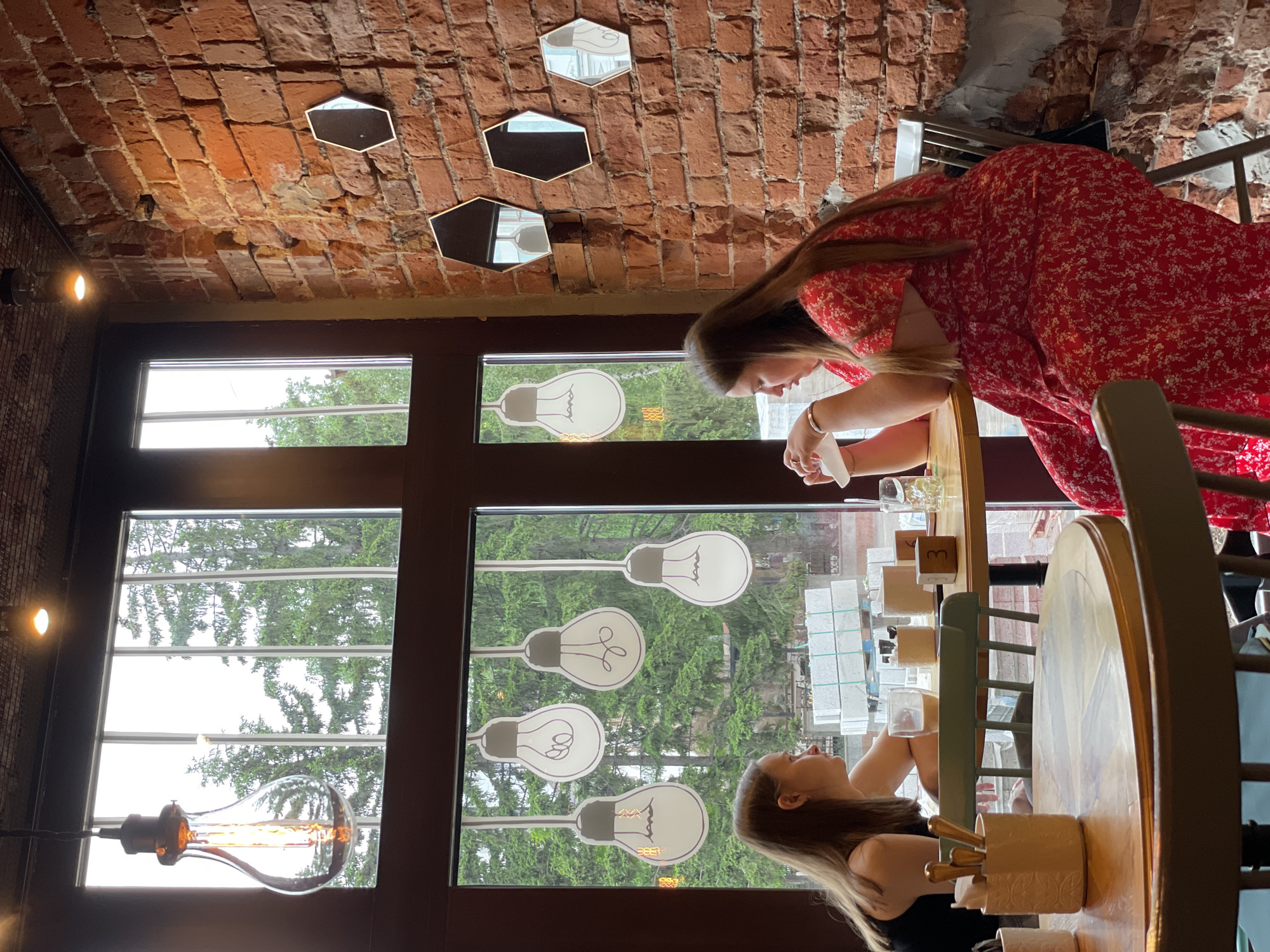 This image depicts the interior of a cafe, likely in Krasnoyarsk, Russia, given the context of the request. Two young women are seated at tables, facing each other, engaged in conversation. The woman on the left, with long brown hair, wears a black top. The woman on the right, also with long brown hair, is dressed in a red floral-patterned dress. They are seated near a large window that offers a view of trees and some construction materials outside.
The cafe's decor features a brick wall with hexagonal mirrors and a prominent Edison-style lightbulb hanging from the ceiling. Inside the window, there are decorative cutouts of lightbulbs. On the tables, there are glasses, napkins, and small decorative cubes with numbers on them, suggesting a table number. One woman is holding a napkin. The environment appears to be a casual dining or coffee shop setting.