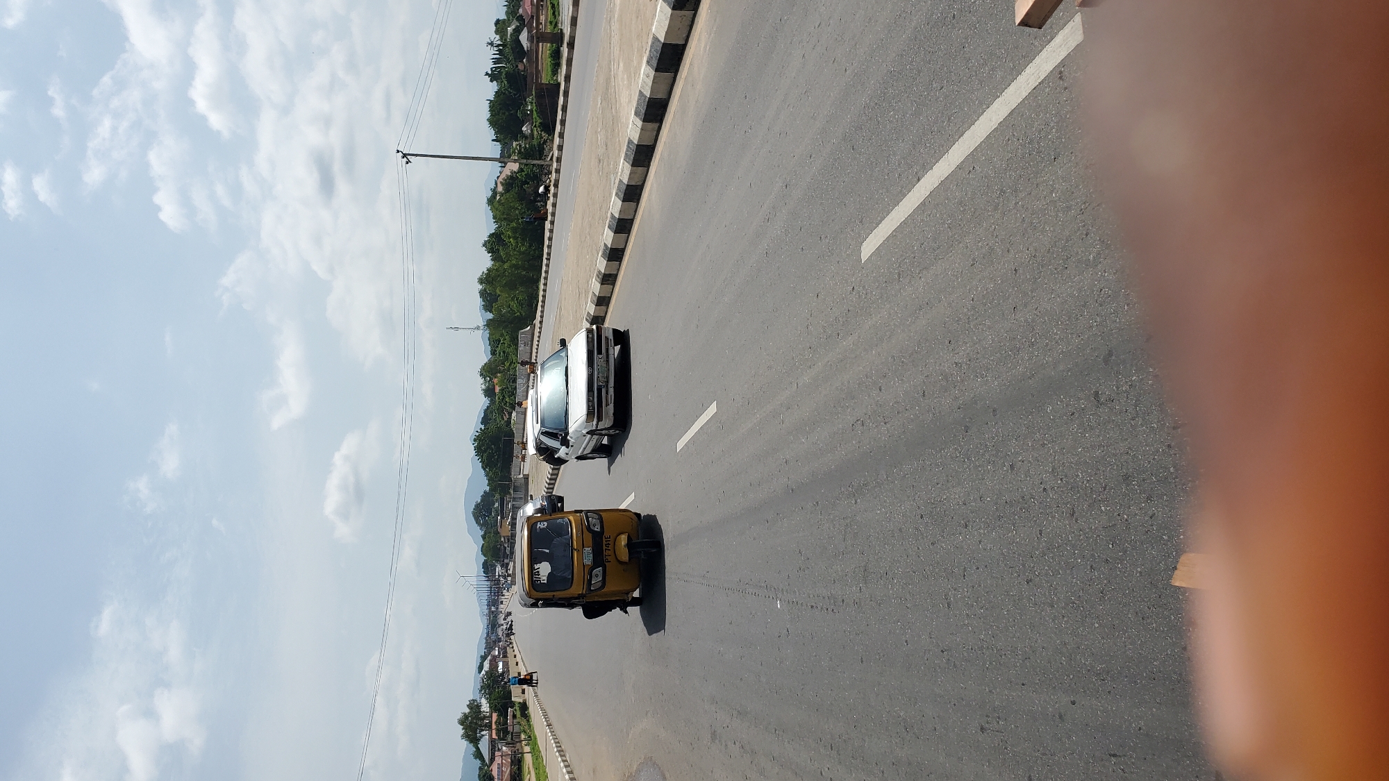 The image shows a street scene in Jalingo, Nigeria, on a bright, overcast day. A yellow tricycle, also known as a Keke Napep, is traveling on the right side of the road. A white sedan is also on the road, to the right of the tricycle. The road is paved and has lane markings. There are trees and buildings visible in the background, as well as some hills in the distance. The sky is cloudy. The image appears to have been taken from a moving vehicle. A blurry orange-colored area, likely a finger or hand, is visible in the foreground, obscuring part of the lower frame.