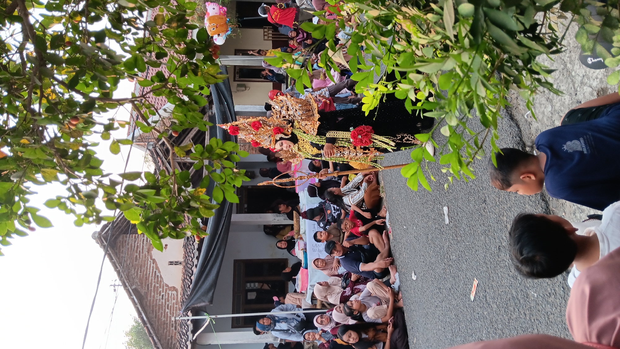 In the city of Cerme, Indonesia, a cultural event is taking place outdoors on a paved street. The central focus is a person dressed in a traditional, elaborate costume, featuring a golden headdress adorned with red flowers and intricate jewelry. This individual holds a wooden staff and is positioned in the foreground, slightly to the right of the image's center.
A crowd of people, predominantly women and children, are seated on the ground on both sides of the street, observing the event. Many are wearing headscarves. Behind the seated spectators, a white tent structure is visible, suggesting a market or exhibition area. A few individuals stand near the tent, and some are engaged in conversation. In the background, traditional Indonesian houses with brick walls and tiled roofs are visible. Overhead, branches of a leafy tree with small reddish fruits partially obscure the sky. Colorful balloons, one shaped like a smiling face, are tied to a pole on the right side of the scene. The overall atmosphere appears festive and communal.