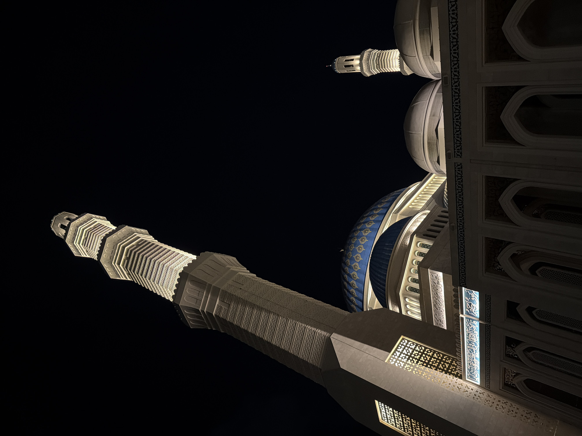 This is a nighttime, low-angle shot of the Hazret Sultan Mosque in Astana, Kazakhstan. The image is dominated by the mosque's architecture, illuminated by artificial lights against a dark, cloudless sky.
In the foreground and to the left, a tall, slender minaret rises at an acute angle, its white facade intricately patterned and lit from within and along its edges. The minaret's base is a solid, octagonal structure. To the right of the minaret, a large, vibrant blue dome, adorned with golden patterns, is visible. Below the dome, the mosque's main structure features arched windows and decorative friezes, with Arabic calligraphy illuminated in a cool blue light. Further to the right, two smaller, lighter-colored domes are partially visible. In the background, a second, smaller minaret with a more traditional bulbous top is also lit. The overall scene is serene and majestic, highlighting the mosque's grandeur under the night sky. There are no people visible in the image.