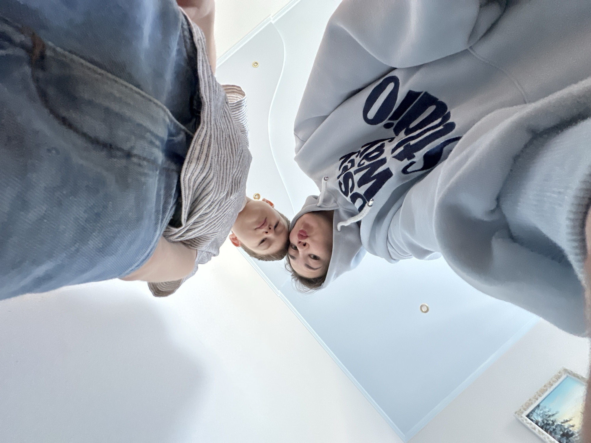 Two people are positioned above the camera in an indoor environment with white walls and a white ceiling. One person wears a light blue hooded sweatshirt with dark lettering, while the other wears denim or a similar blue fabric and a striped garment beneath. The scene suggests a selfie or a photo taken at a low angle, looking up at the subjects. In the background, there is a framed picture on the wall. The location is unclear and therefore not possible to confirm it is in Ilyinsky, Russia.