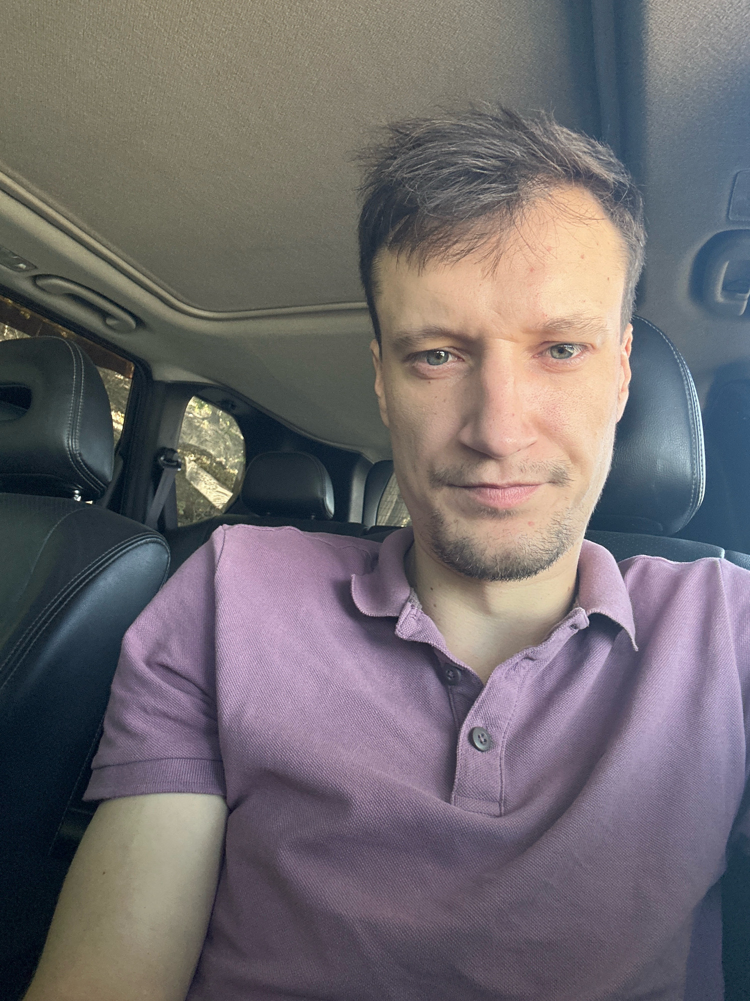 A man is sitting in the driver's seat of a car with a sunroof open. He is wearing a light purple polo shirt. The car appears to be parked outdoors, with trees visible through the windows. The image is a selfie, taken from the man's perspective. The location is Makiv, Ukraine.