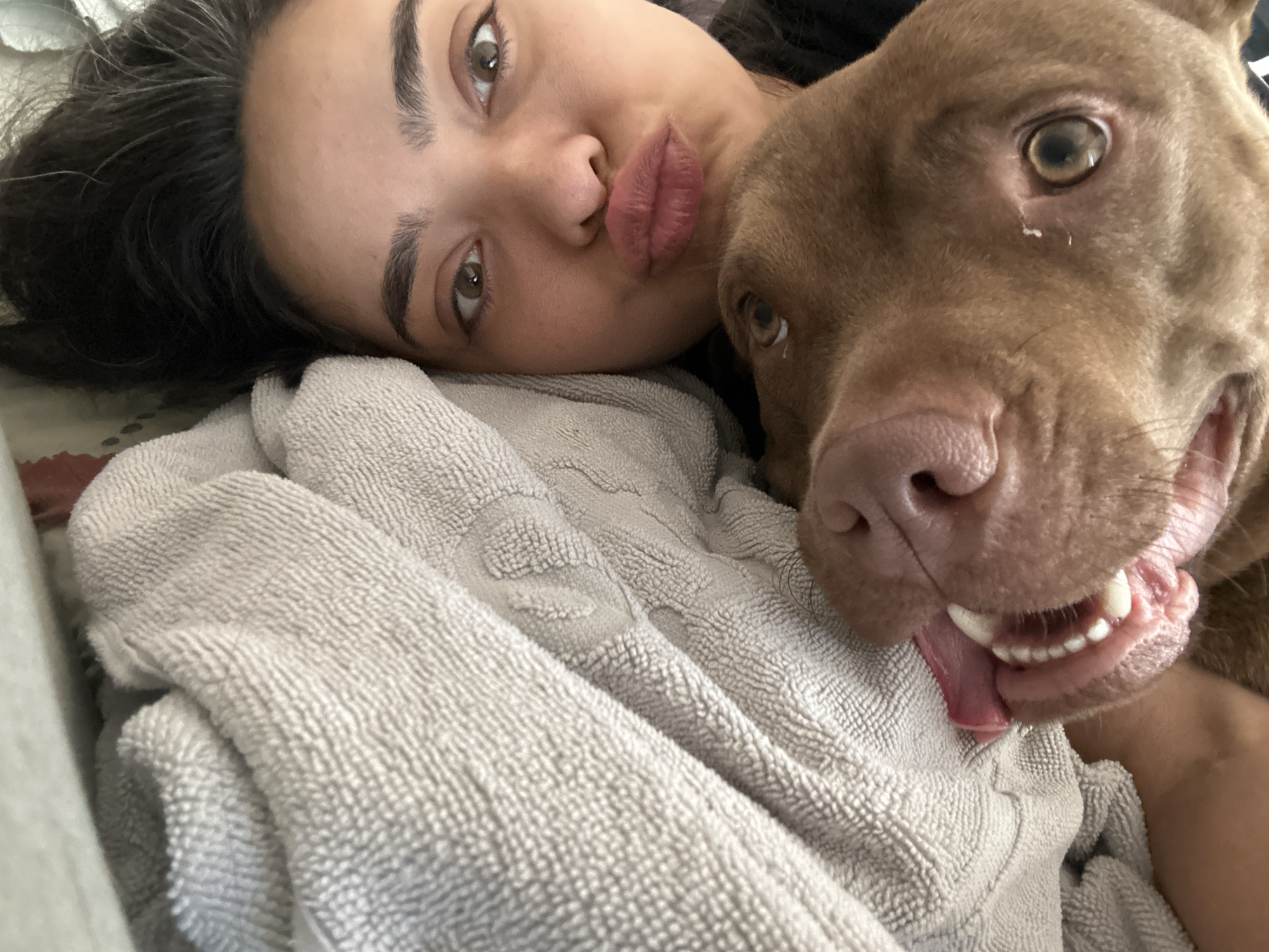 A close-up selfie shows a young woman and a brown dog lying together on a grey towel. The woman is on the left, with her head resting on the towel. She has dark hair and light brown eyes, and her lips are pursed. The dog is on the right, with its head next to the woman's face. It has brown fur and its mouth is open, with its tongue sticking out. The image appears to have been taken indoors, possibly in a bedroom, as indicated by the presence of the towel and the soft lighting. There is no discernible information to indicate the location in the city of Malevo, Bulgaria.