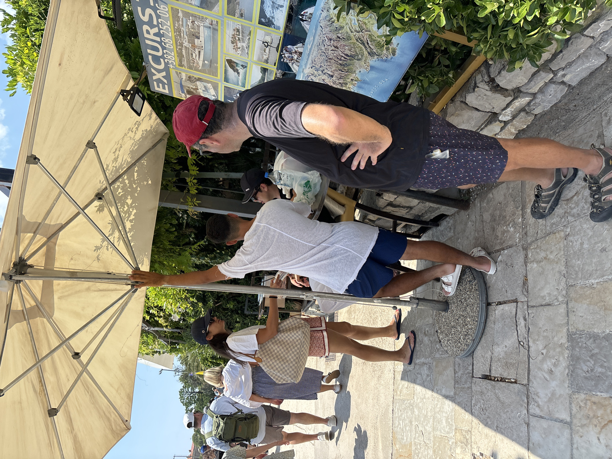 The image captures a sunny outdoor scene in Budva, Montenegro, likely near a tour operator's booth. In the foreground, a person wearing a red cap and a black t-shirt with patterned shorts stands with their hand on their hip, facing right. To their left, a person in a white t-shirt and blue shorts stands looking towards the left. Between them, a person with a black cap and sunglasses, wearing a white t-shirt and patterned shorts, holds a large checkered tote bag. Behind them, a person in a white shirt and plaid skirt walks away, and a person with a green backpack and white shirt walks in the opposite direction.
The setting appears to be a paved area with a large, cream-colored umbrella providing shade. Behind the umbrella, a poster board displays images of boats and a map, advertising excursions. Green foliage and a stone wall are visible in the background, contributing to a warm, Mediterranean ambiance. The overall impression is one of a tourist area on a pleasant day.