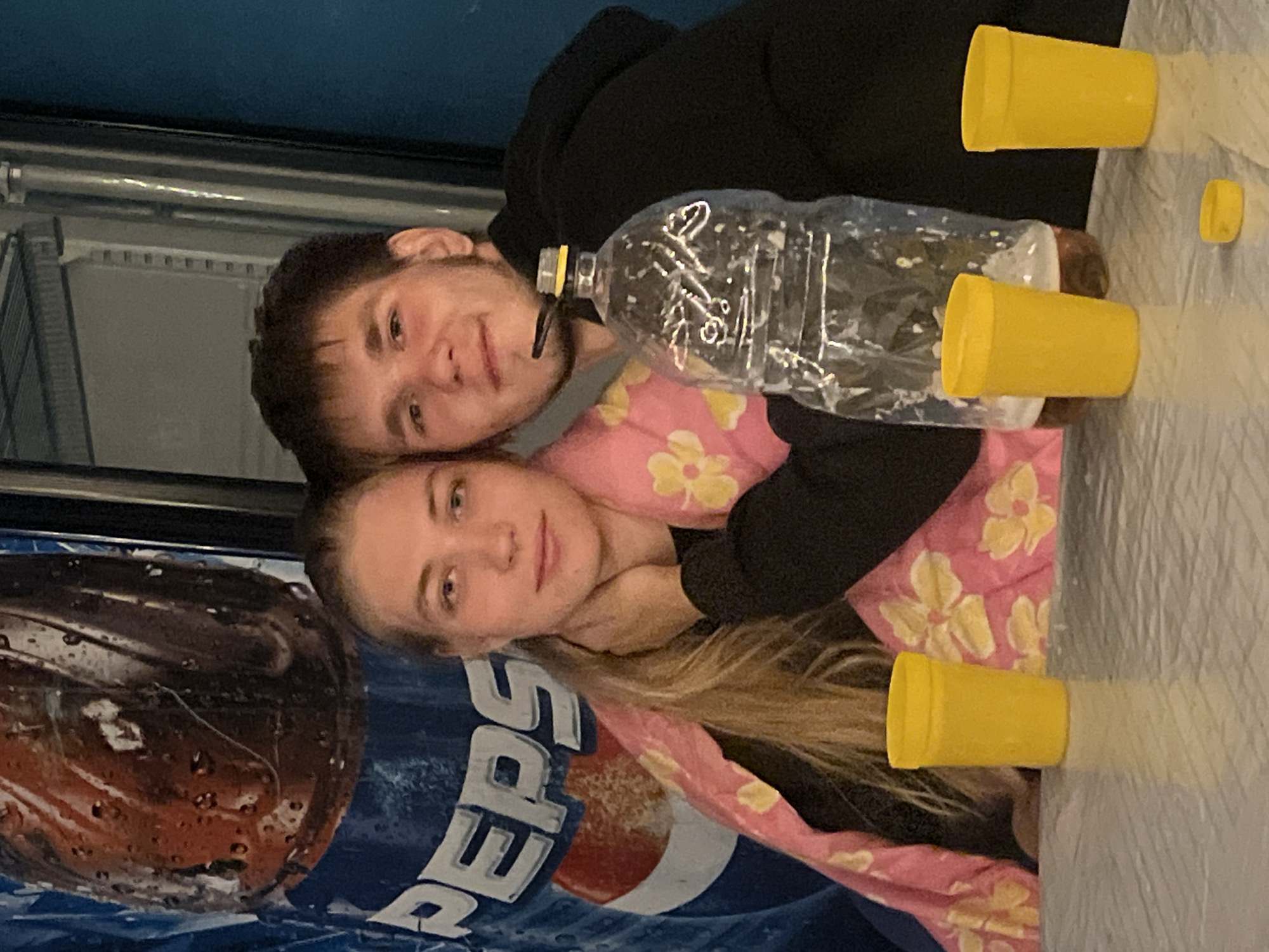 A young couple is posing for a photo in front of a Pepsi advertisement, likely in a public establishment such as a cafe or restaurant in Баянауыл ауылдық округі, Kazakhstan. The woman is on the left, her head resting on the man's shoulder. She has long blonde hair and is wearing a black sweatshirt and a pink blanket with yellow floral patterns. The man is on the right, wearing a black hoodie. They are both looking at the camera with smiles. On the table in front of them are three yellow plastic cups and a large, partially full plastic bottle with a yellow cap. The background features a large Pepsi advertisement, indicating a commercial setting. The lighting appears to be artificial, suggesting an indoor environment.