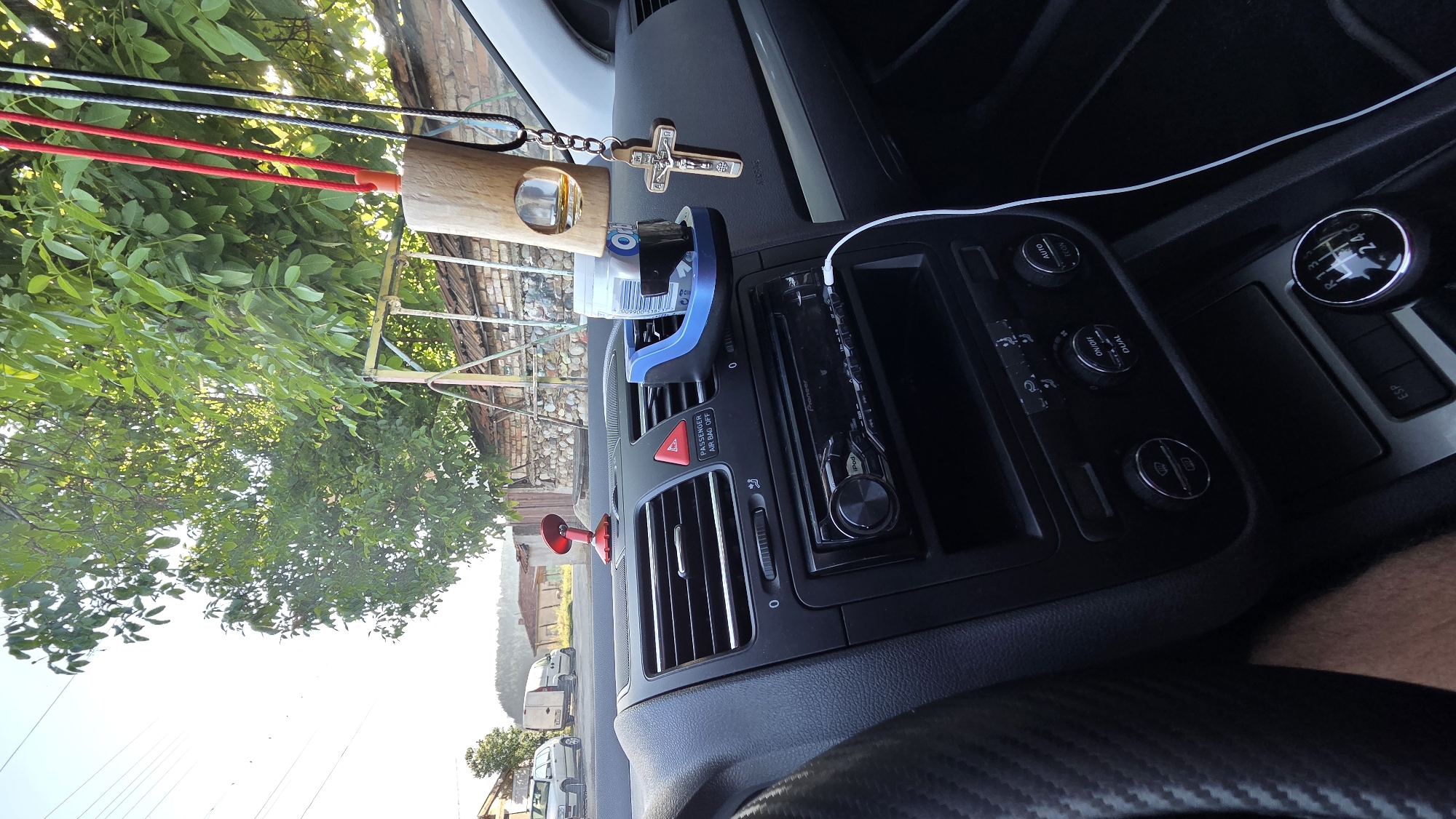 A close-up, interior shot of a car's dashboard shows a silver and black Pioneer car stereo system. Above the stereo are air vents and a red hazard light button with "PASSENGER AIR BAG OFF" text. To the right of the vents, a blue car cup holder is mounted, containing a cylindrical object and what appears to be a clear container with liquid. Hanging from the rearview mirror or a similar fixture above the cup holder are a wooden air freshener with a hole, a crucifix pendant, and two cords, one red and one black. The view extends to the car's gear shift and a portion of the steering wheel on the left. Outside the car, through the windshield, a tree with green leaves is visible against a bright sky, along with houses and a brick wall.